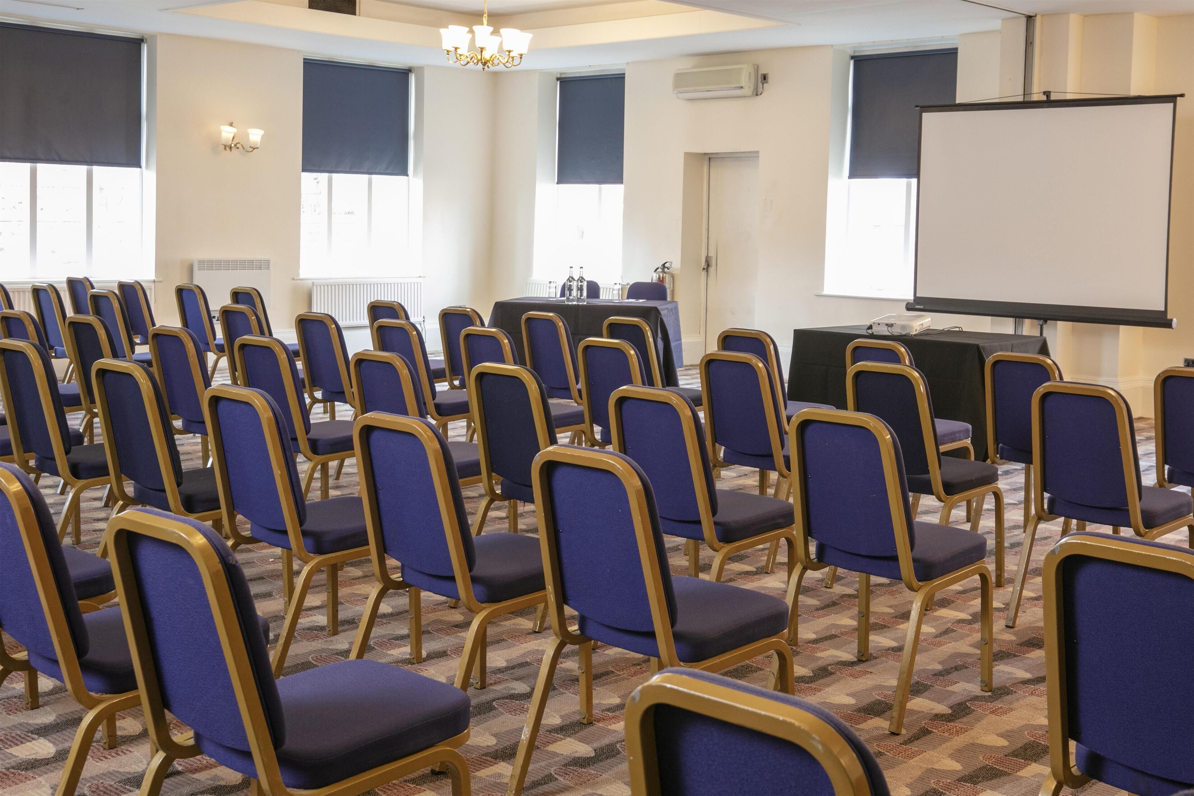 A conference room with rows of blue chairs, a projector screen, and a table with water bottles.