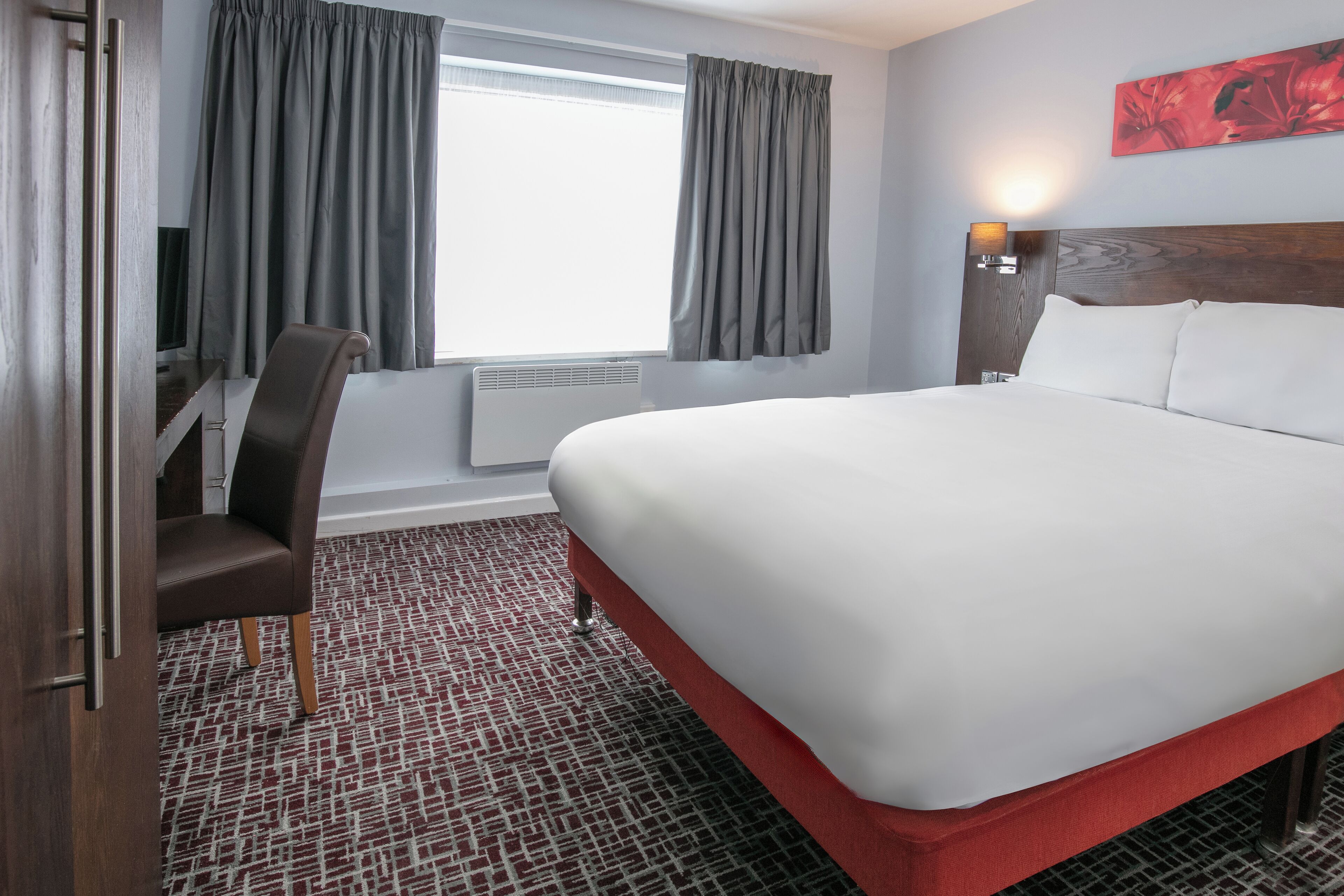 A hotel room with a large bed, a chair, a desk, a window with curtains, and a wall-mounted lamp.