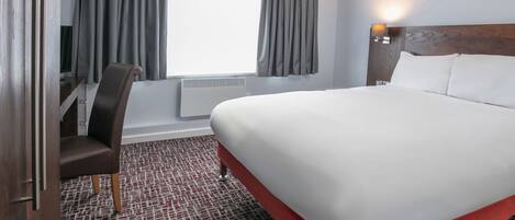 A hotel room with a large bed, a chair, a desk, a window with curtains, and a wall-mounted lamp.