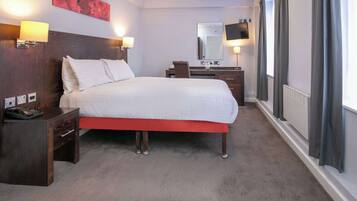 A hotel room with a bed, bedside tables, a desk, a chair, a mirror, a TV, and a window with curtains.