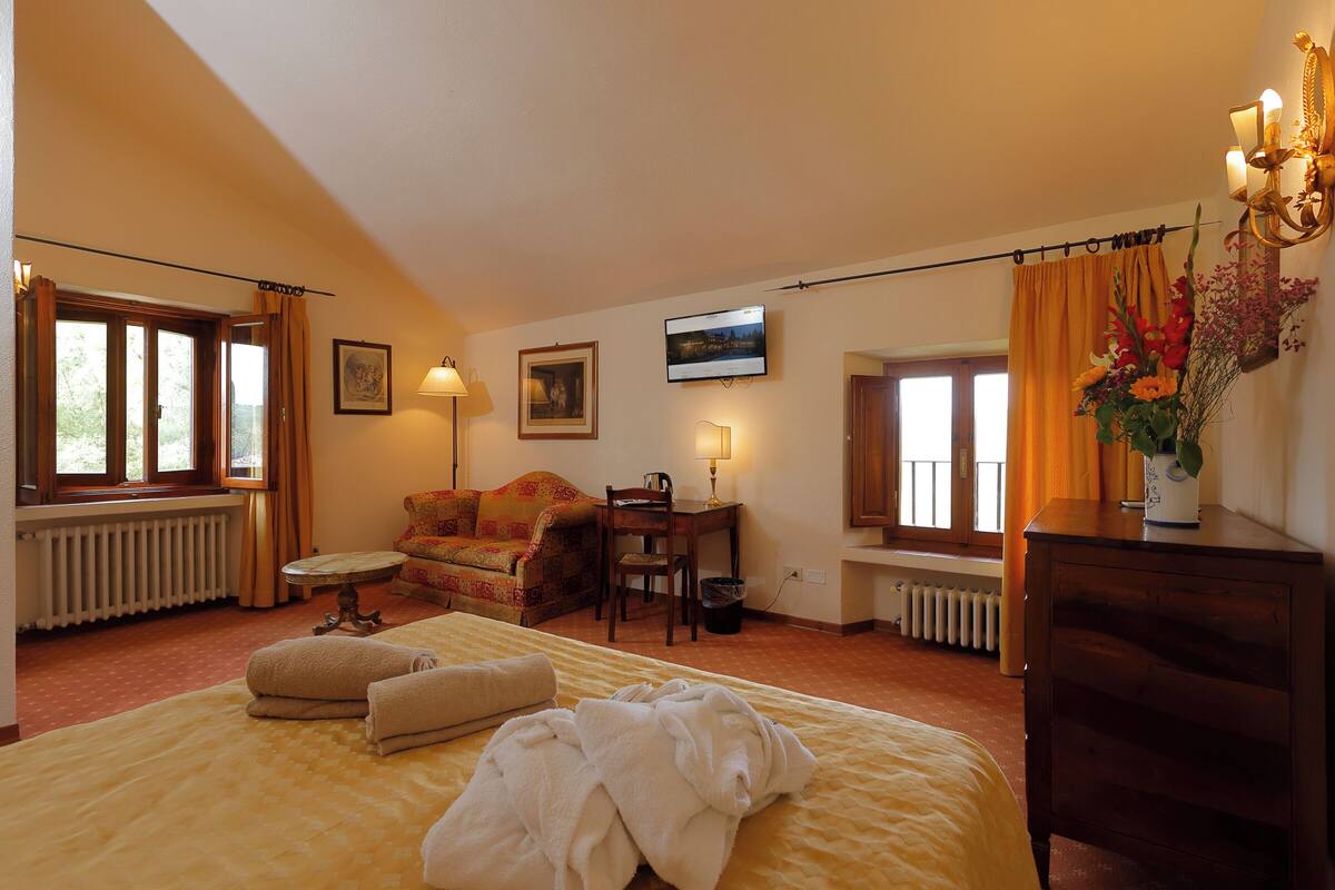 A hotel room with a bed, a sofa, a desk, and a chair. There are windows with curtains, a radiator, and a wall-mounted TV.