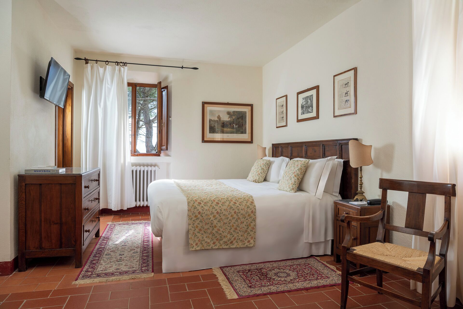 A bedroom with a bed, a wooden chair, a desk with a television, and a window with curtains.