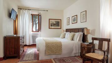 A bedroom with a bed, a wooden chair, a desk with a television, and a window with curtains.