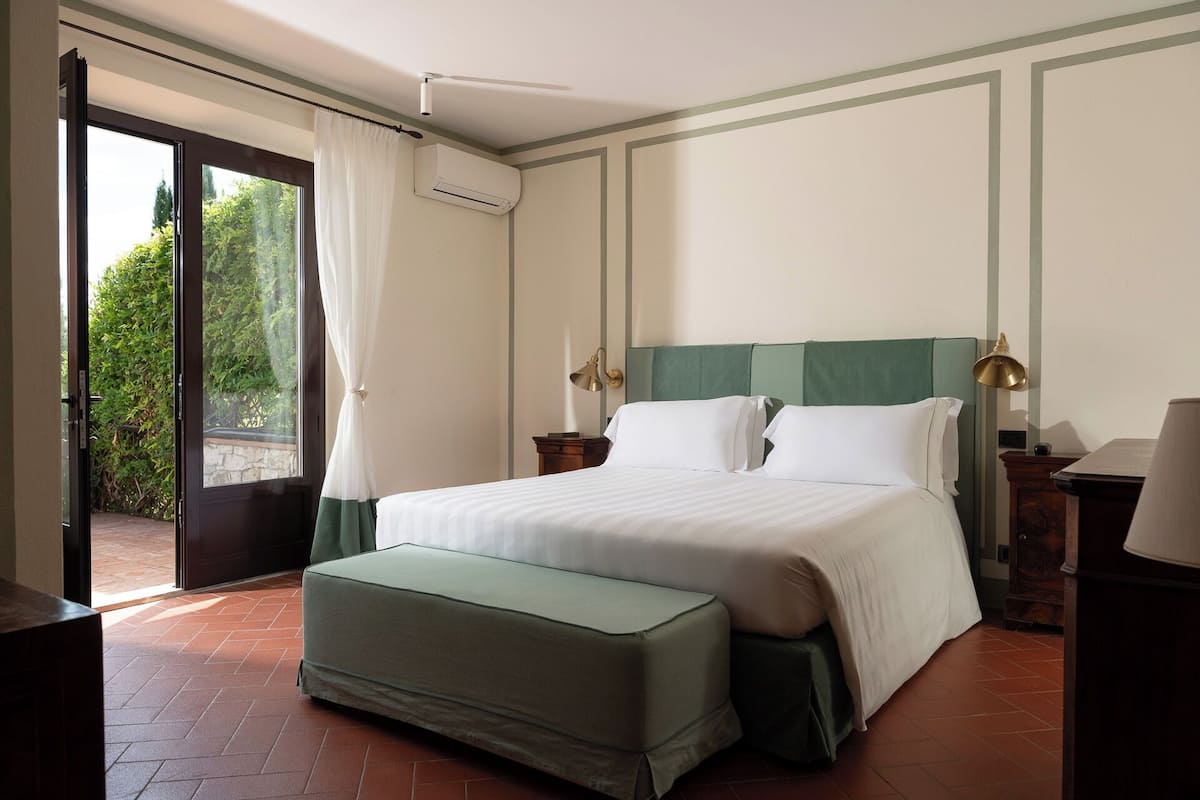 A hotel room with a bed, an ottoman, a nightstand, a lamp, and an air conditioning unit.