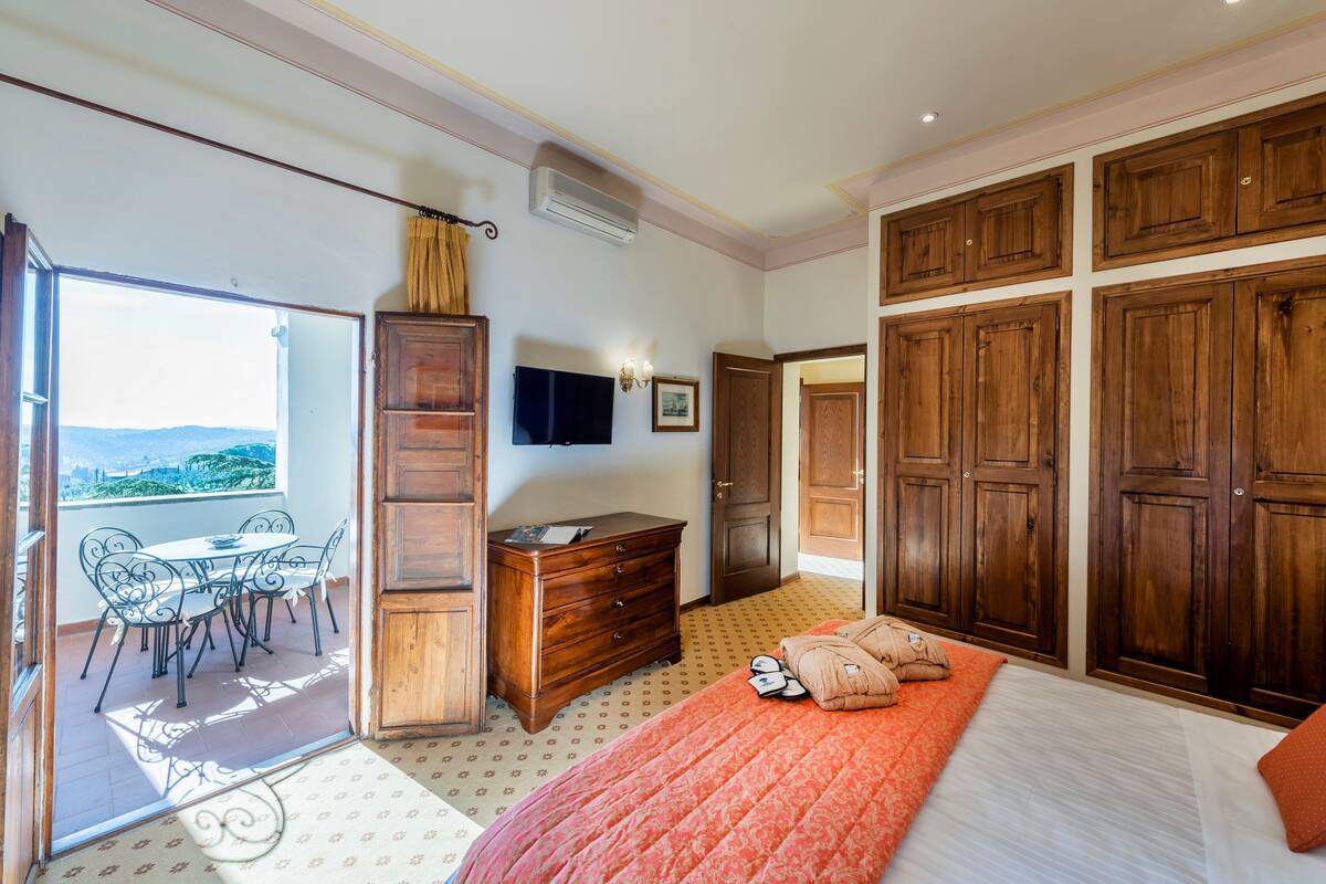 A bedroom with a bed, wooden wardrobe, a desk, a TV, and a balcony with a table and chairs.