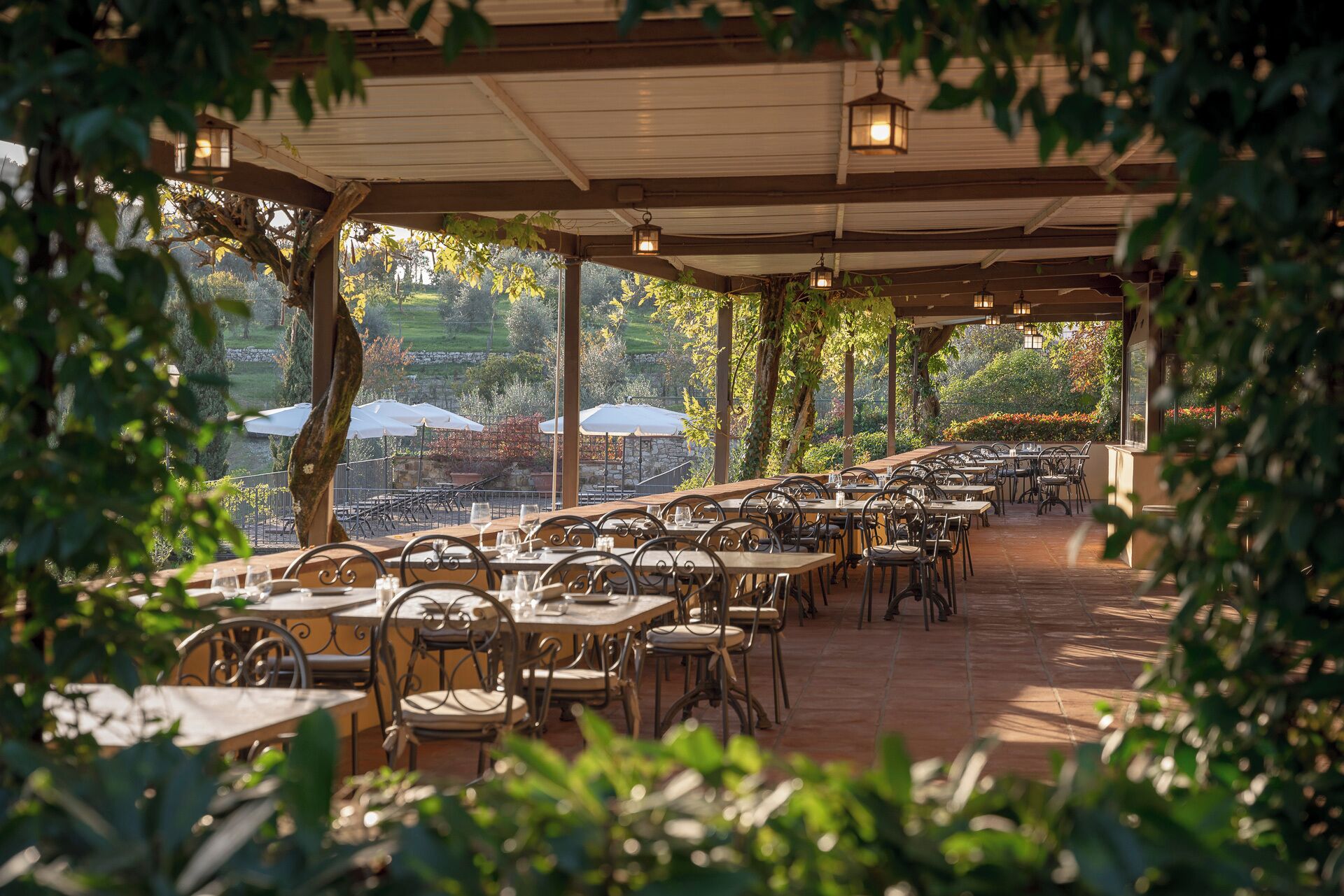 An outdoor dining area with tables and chairs, covered by a roof, and surrounded by greenery.