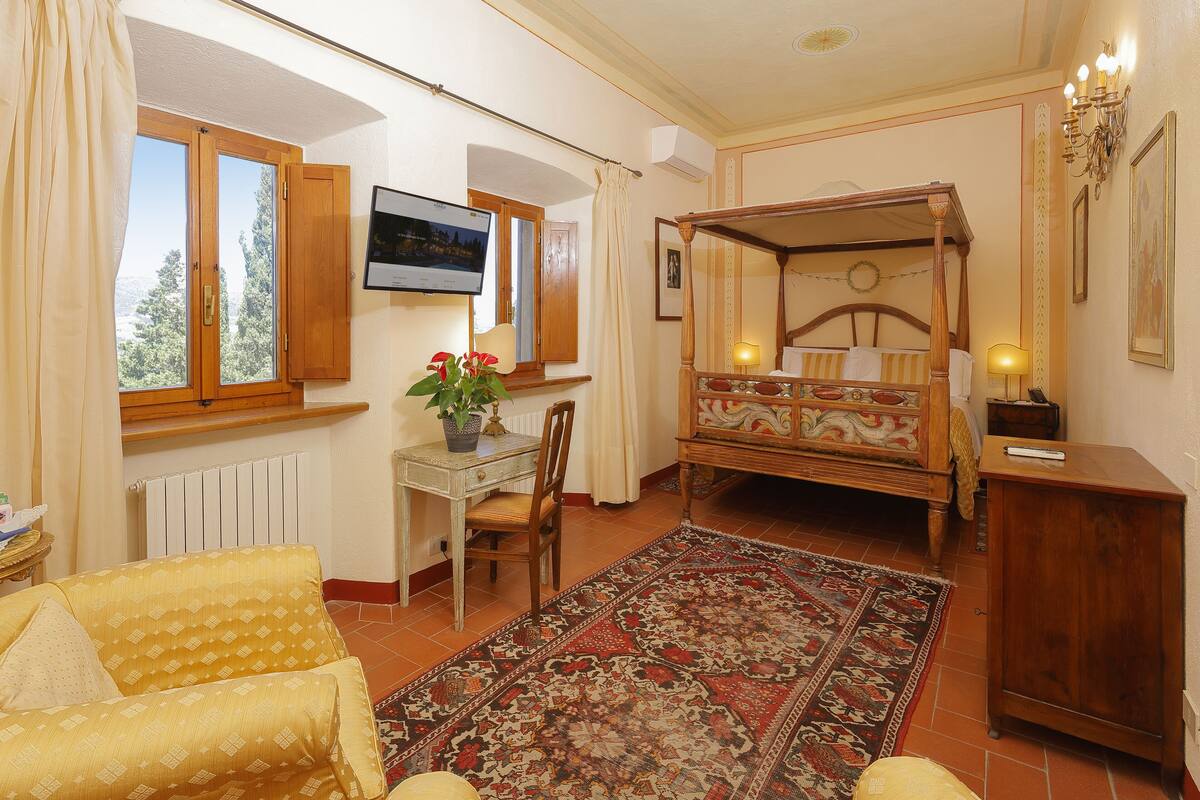 A room with a four-poster bed, a desk with a computer, a chair, a large rug, and a window with curtains.