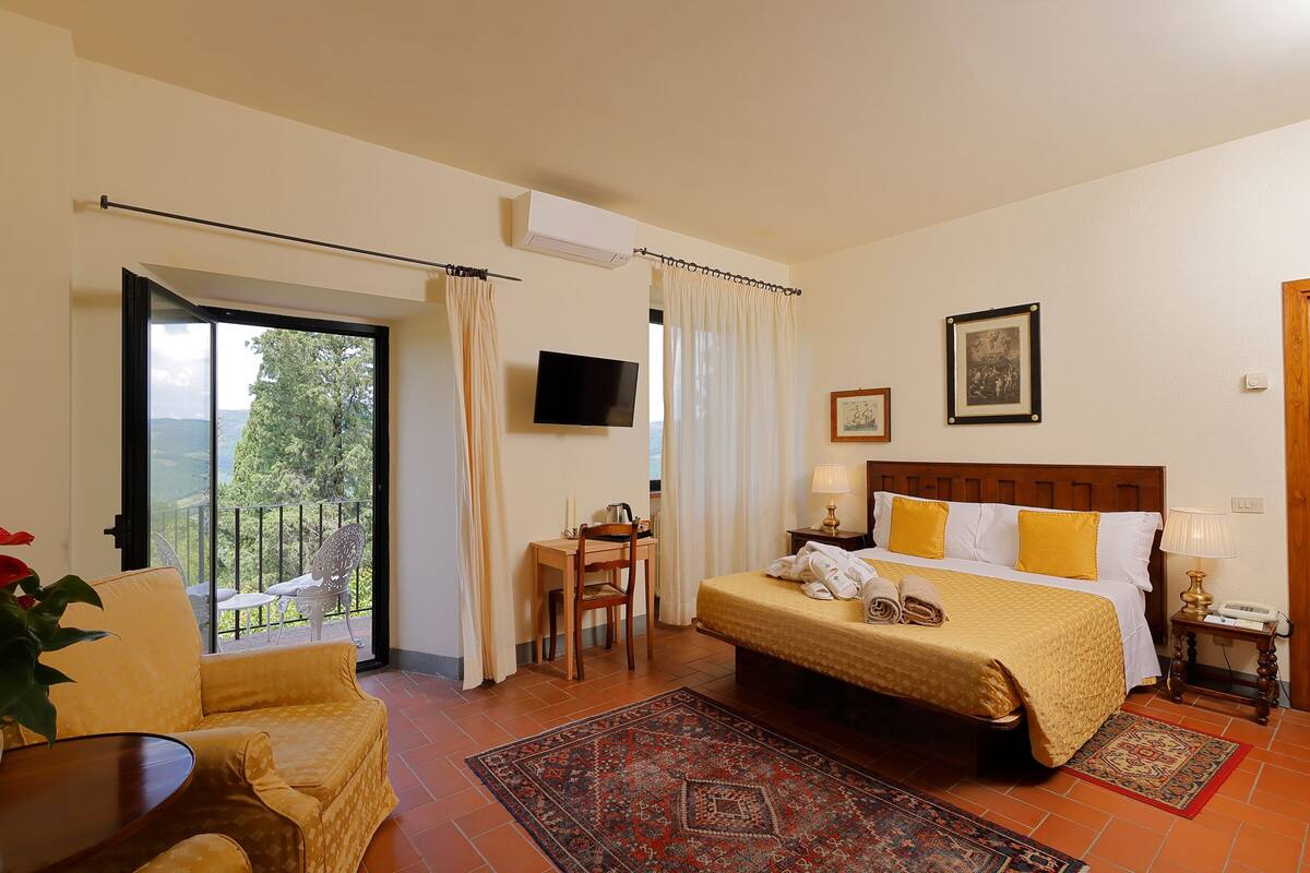 A hotel room with a bed, a desk, a chair, a TV, and a balcony with a view of greenery.