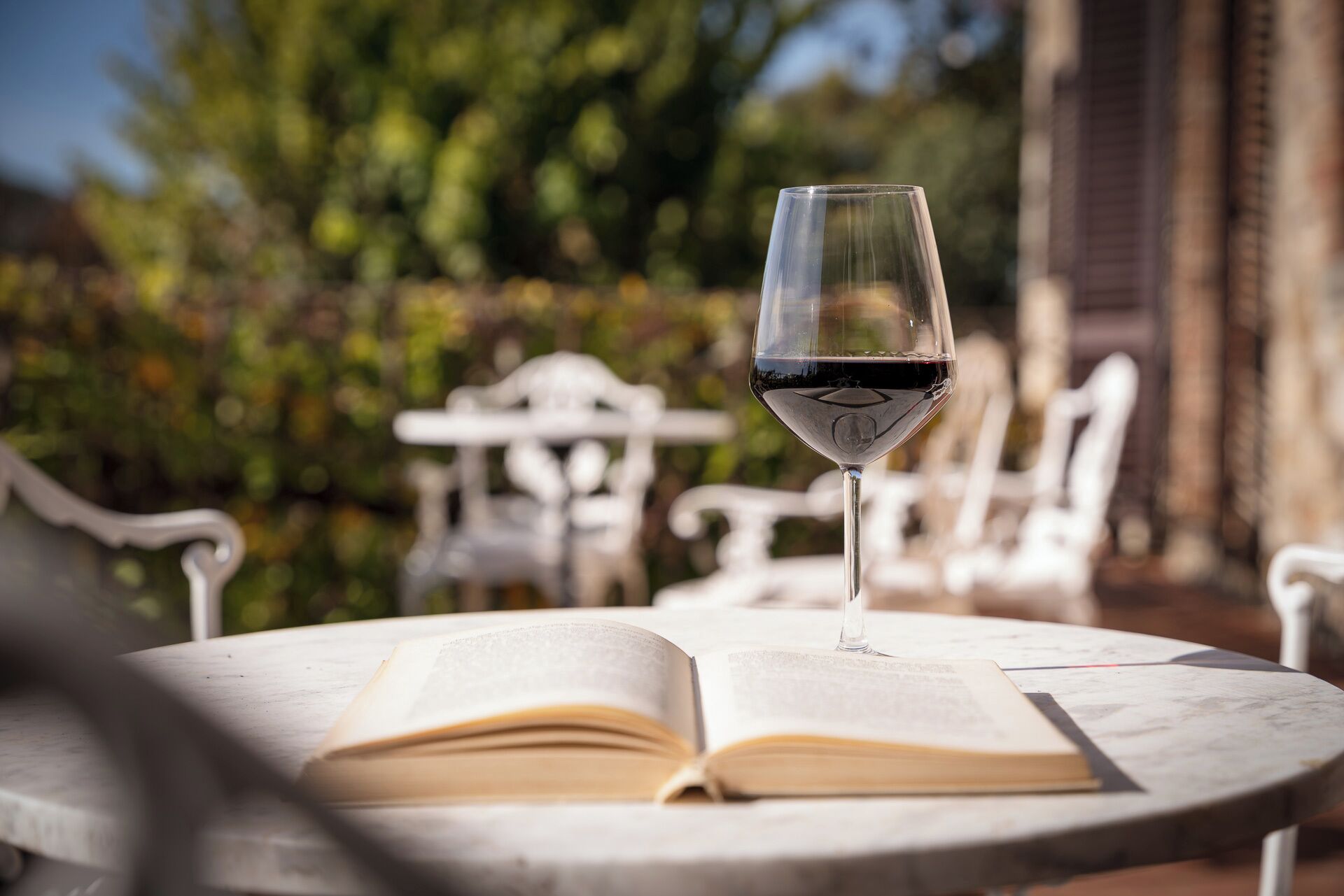 An open book on a round table with a glass of red wine, set outdoors with white chairs and greenery in the background.