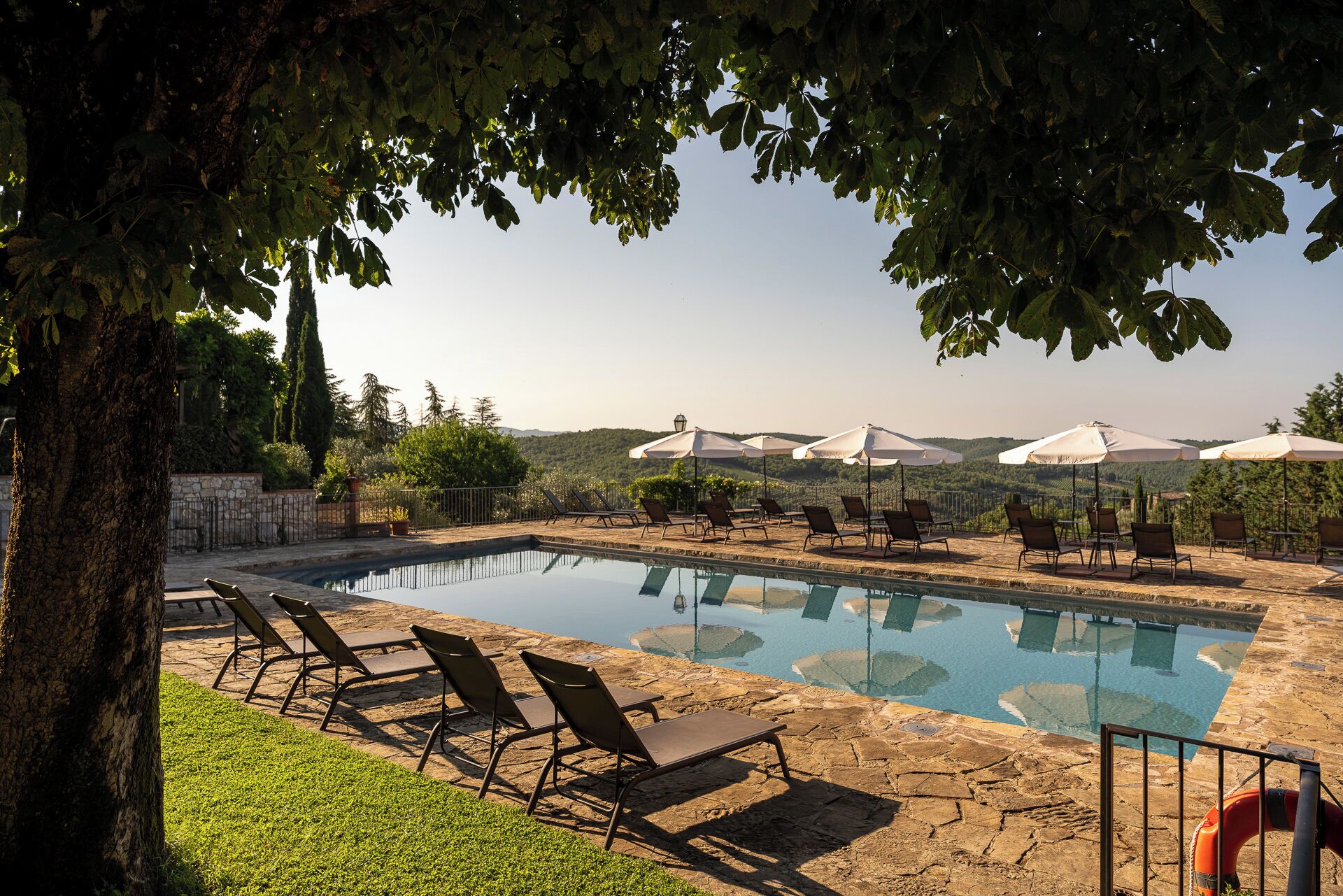 A pool area with lounge chairs and umbrellas, surrounded by trees and a view of hills.