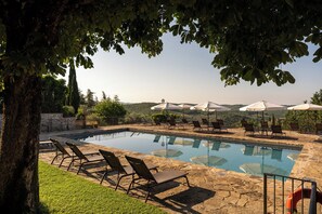 A pool area with lounge chairs and umbrellas, surrounded by trees and a view of hills.