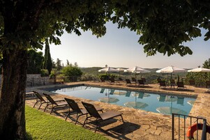 Un área de piscina con sillones y sombrillas, rodeada de árboles y vistas a colinas.