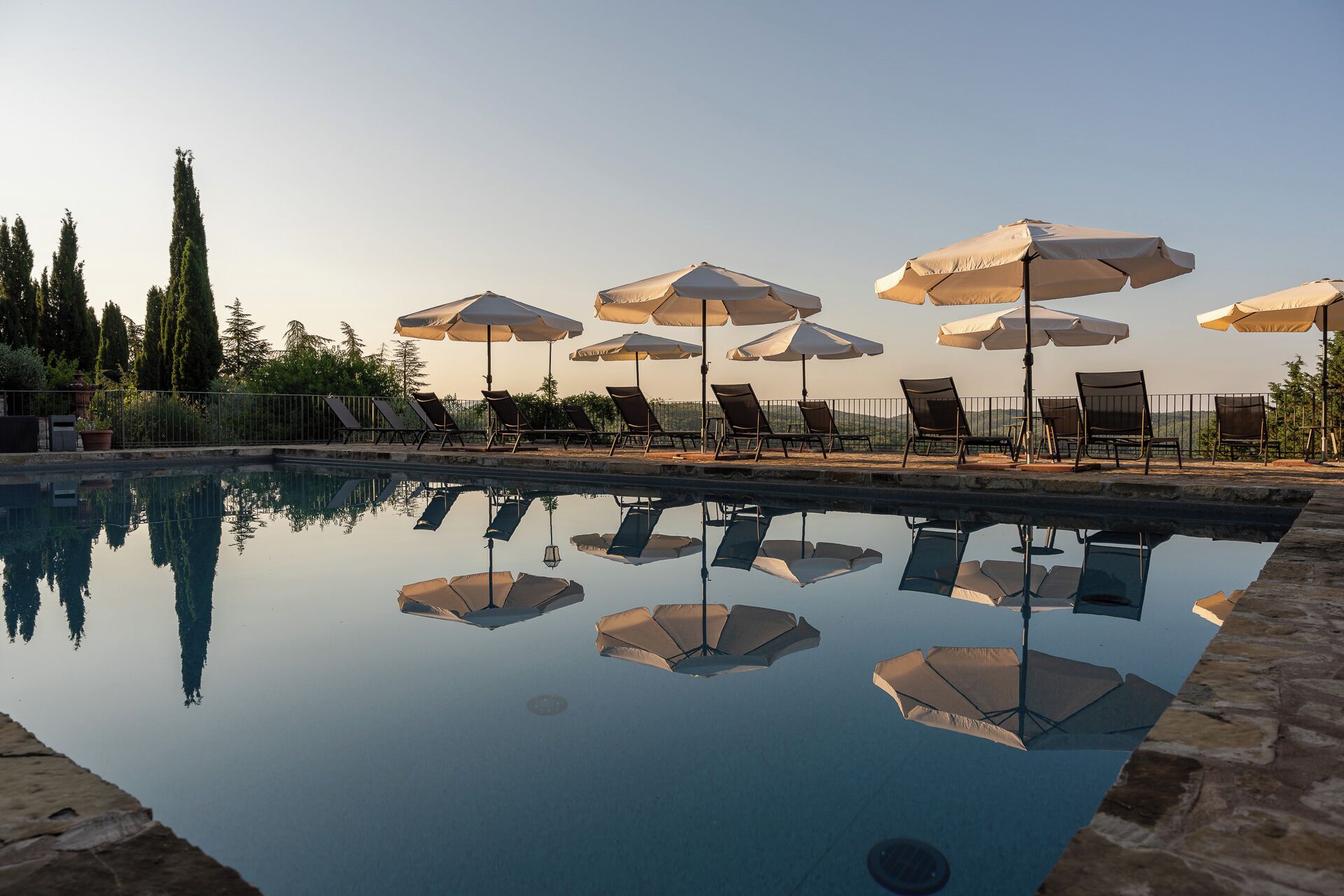Zona de piscina con múltiples sillas de descanso y sombrillas, rodeada por una valla y árboles.