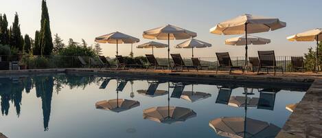 Zona de piscina con múltiples sillas de descanso y sombrillas, rodeada por una valla y árboles.