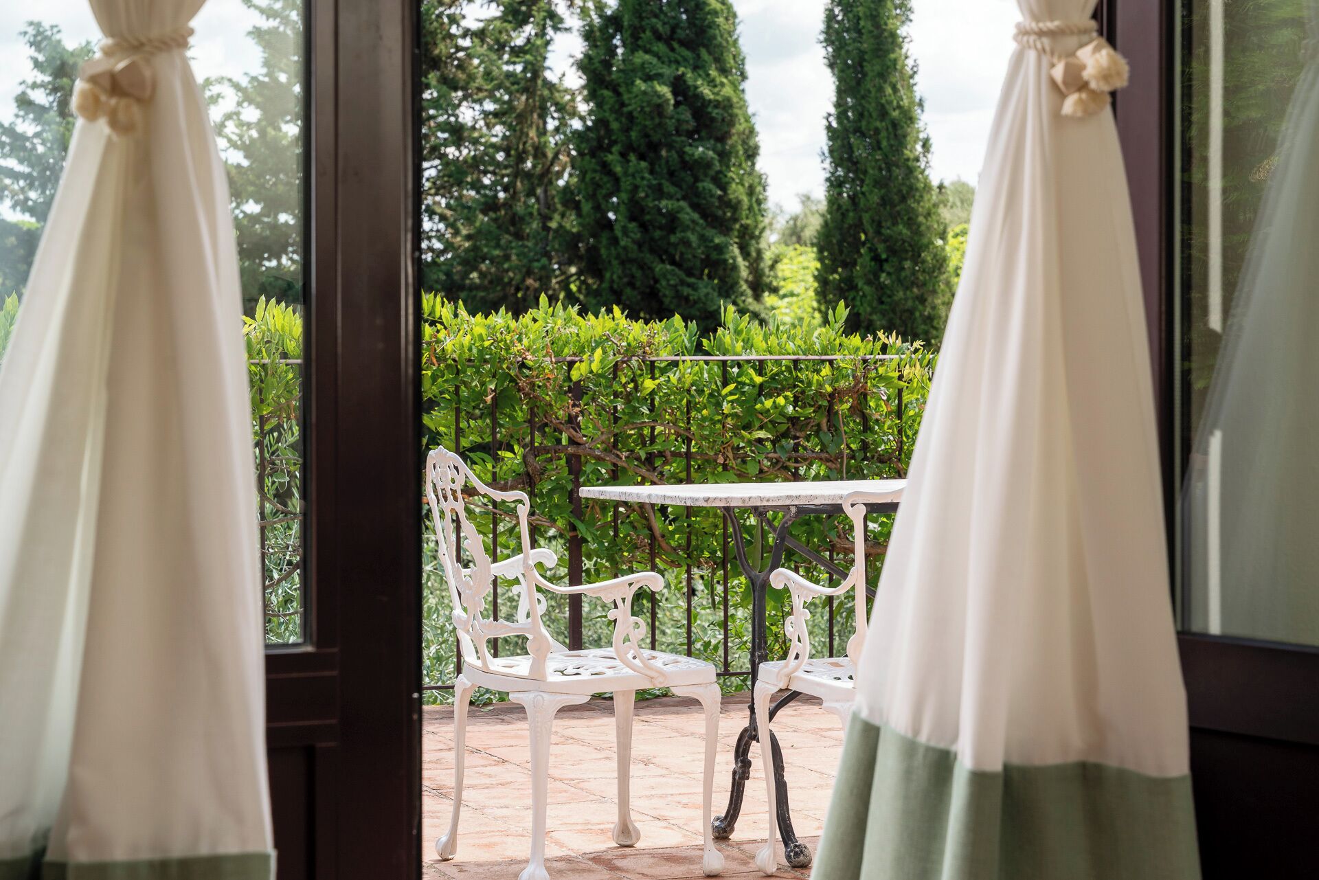 A view through a window with white curtains, featuring a white outdoor table and chairs, a hedge, and tall trees in the background.