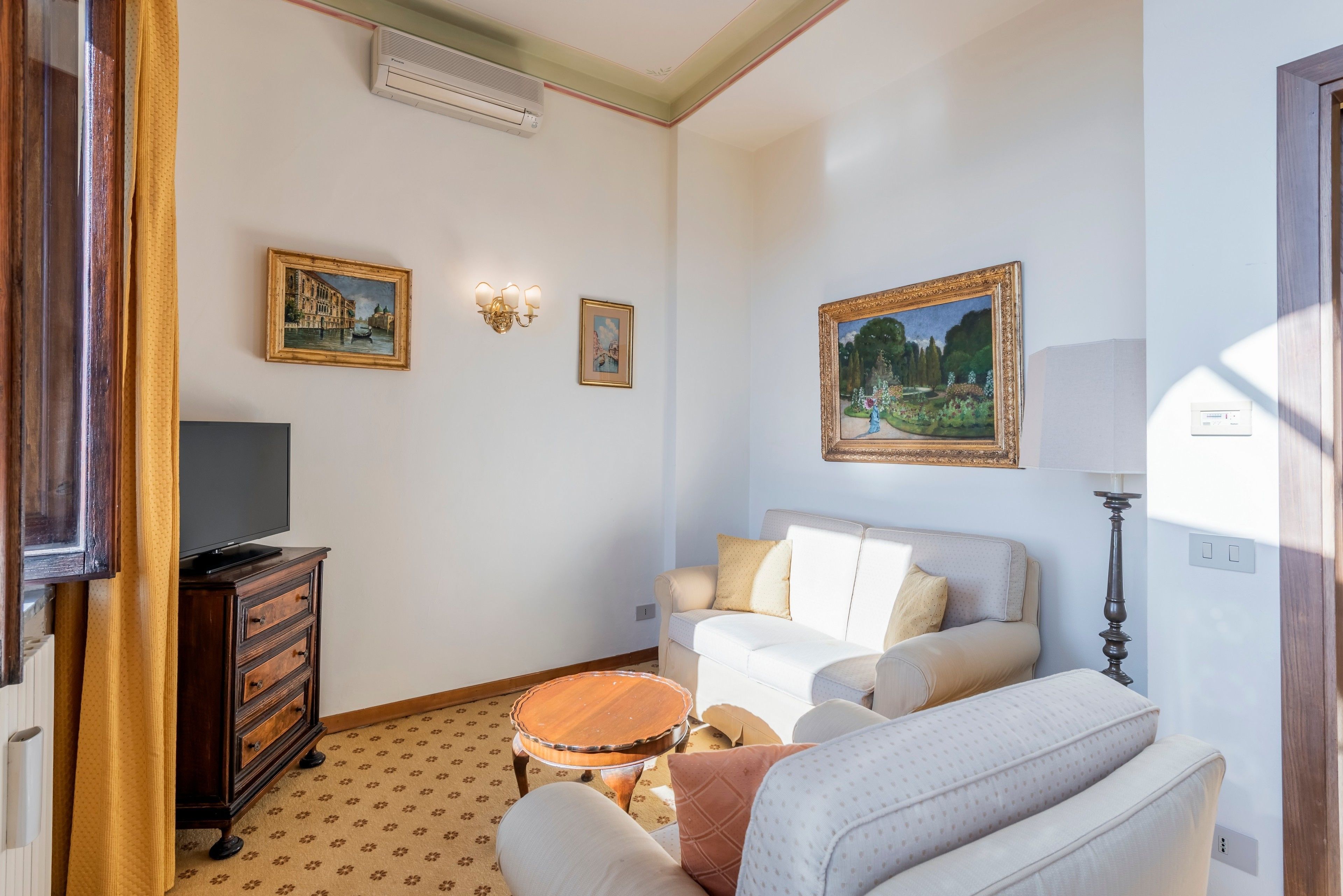 A living room with a television on a wooden cabinet, a white sofa, a patterned rug, and two framed paintings on the wall.