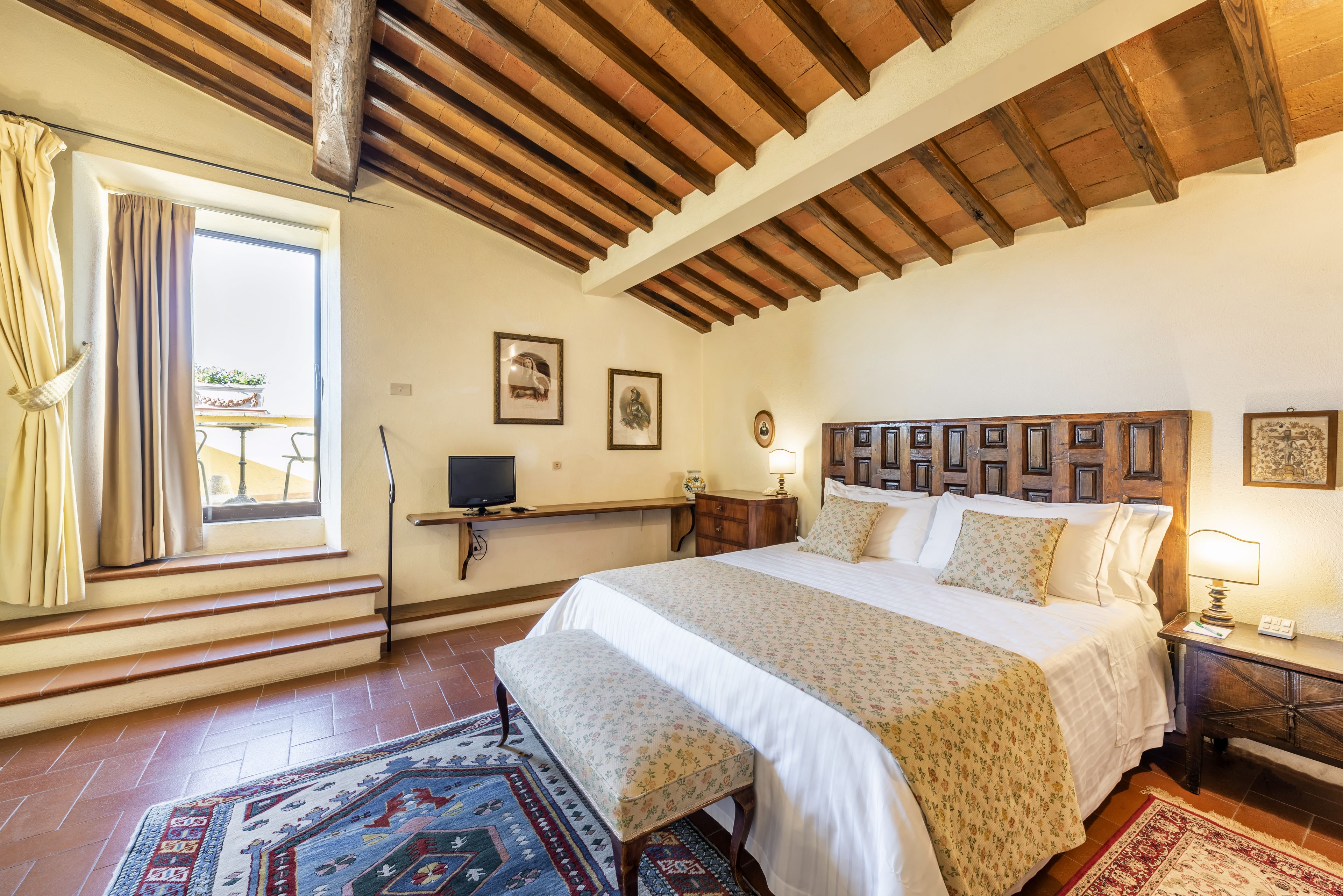 A bedroom with a large bed, wooden headboard, a bench, a TV, and a window with curtains.