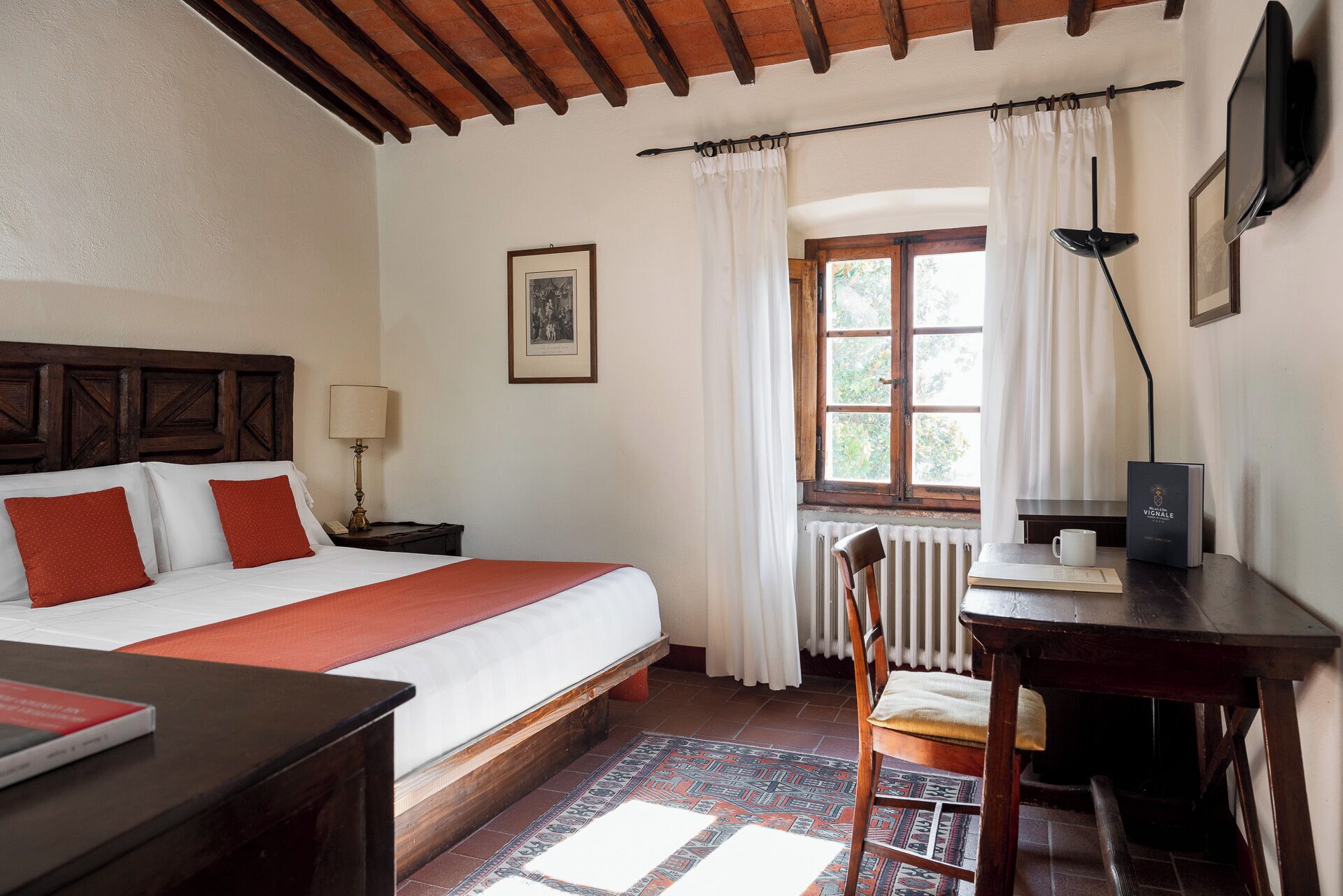 A hotel room with a bed, a desk, a chair, a window with curtains, a radiator, and a framed picture on the wall.