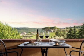 A table with a bottle of wine, two glasses, a plate with food, and a bowl on a balcony overlooking a scenic landscape.