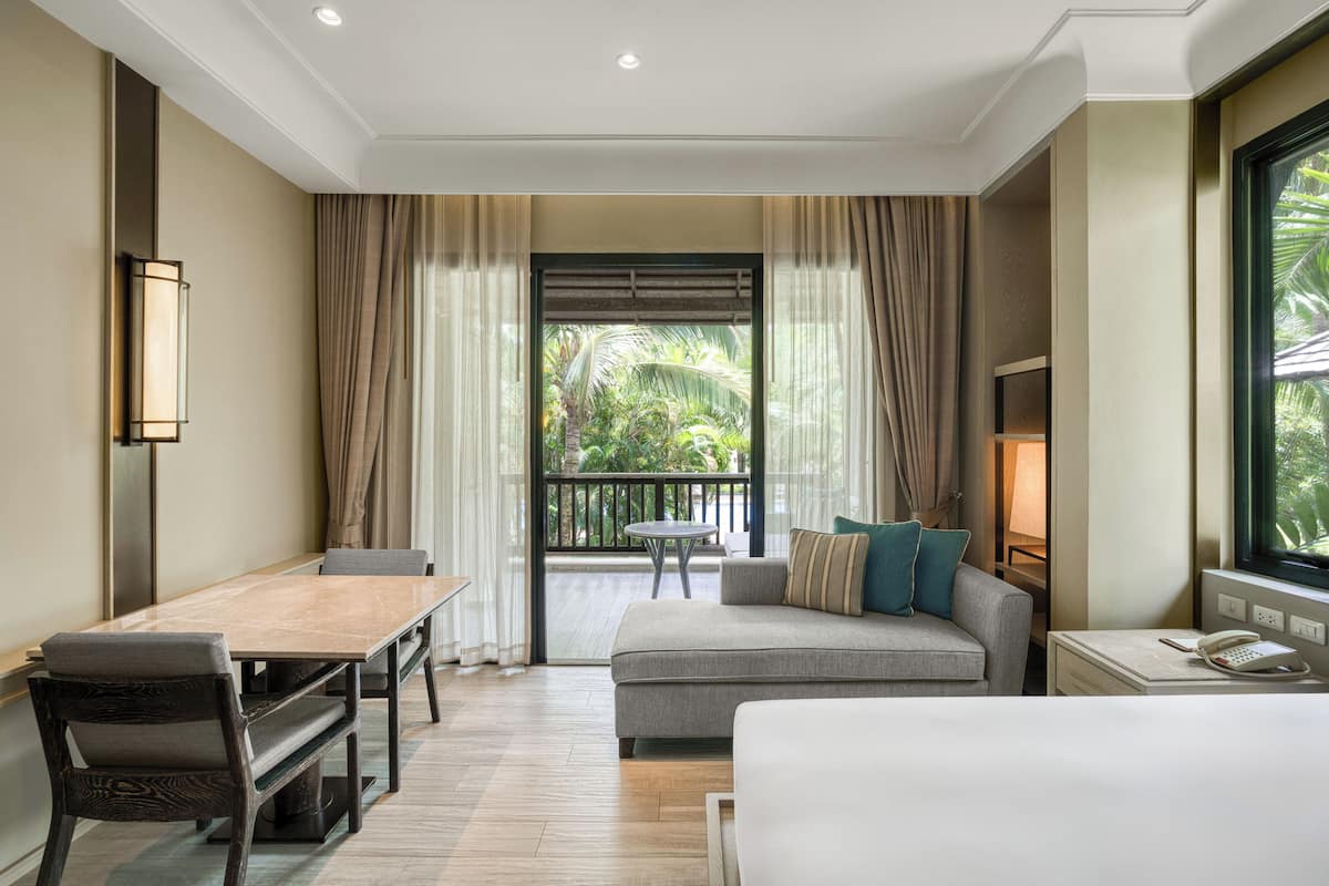 A hotel room with a bed, a sofa, a dining table, and a balcony with a view of greenery.