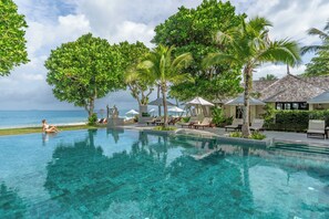A clear pool with lounge chairs and umbrellas, surrounded by palm trees and a view of the ocean.