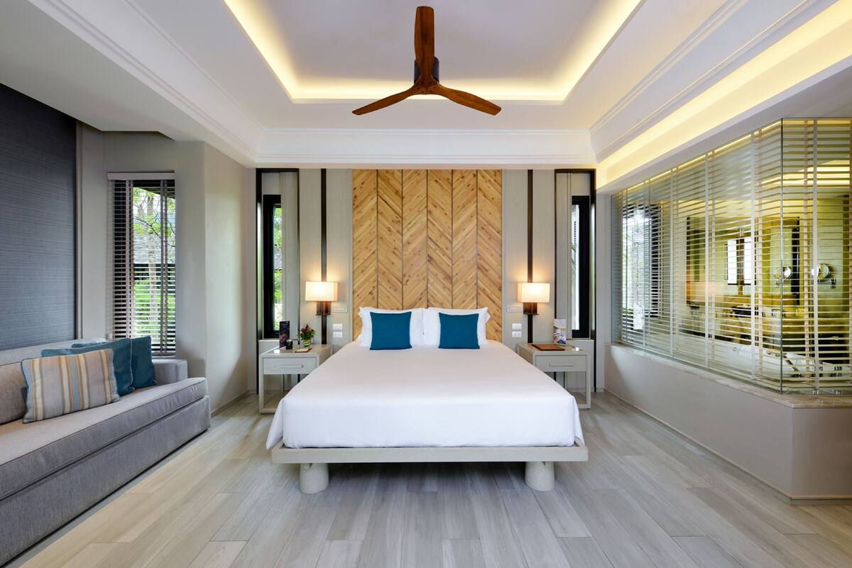 A modern bedroom with a large bed, a wooden headboard, bedside tables with lamps, a sofa, and a view through the window.