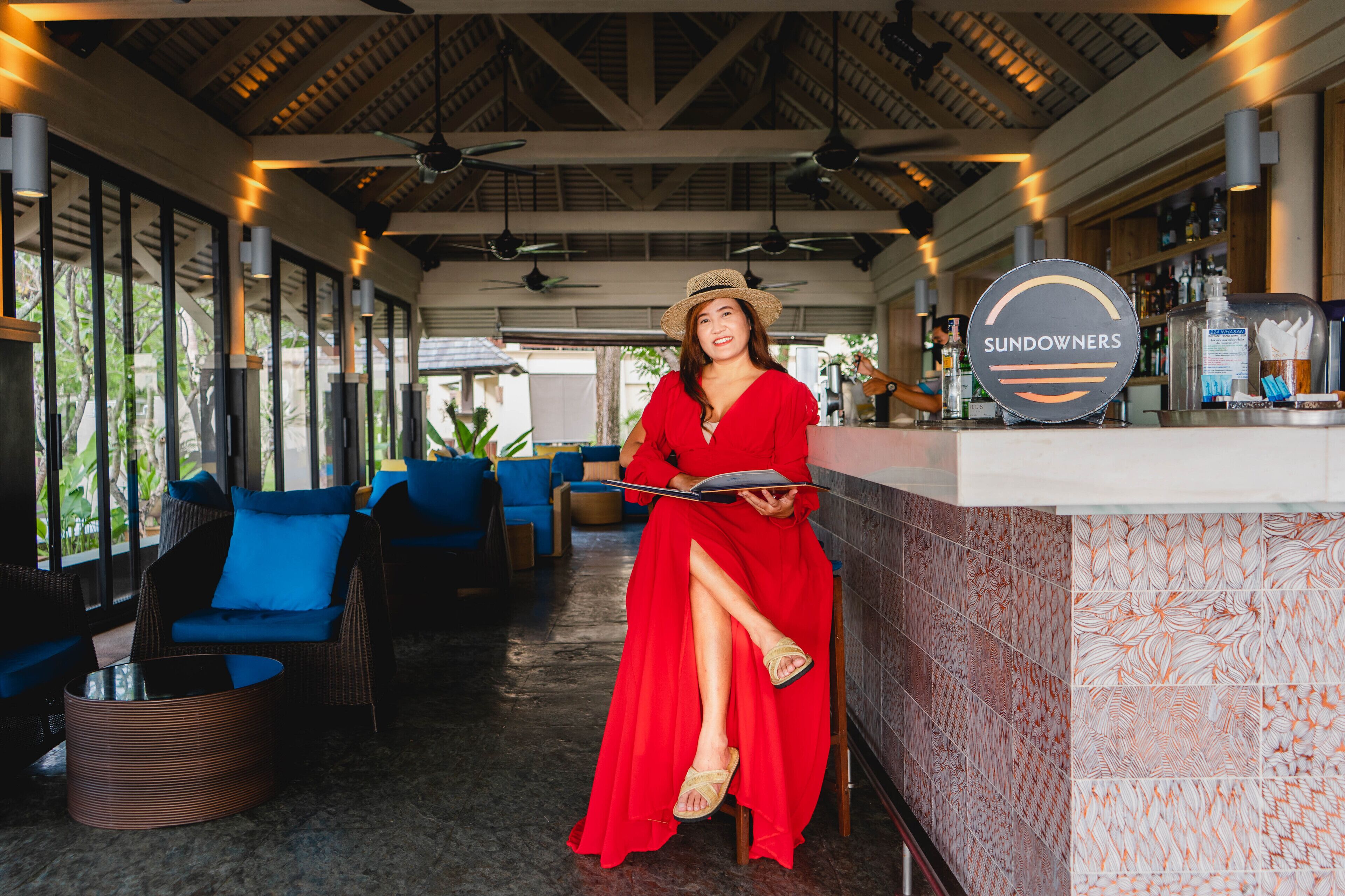 A woman in a red dress sitting at a bar with a sign reading "SUNDOWNERS".