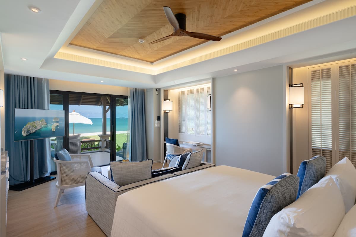 A modern hotel room with a large bed, a ceiling fan, a TV, and a balcony with a view of the ocean.