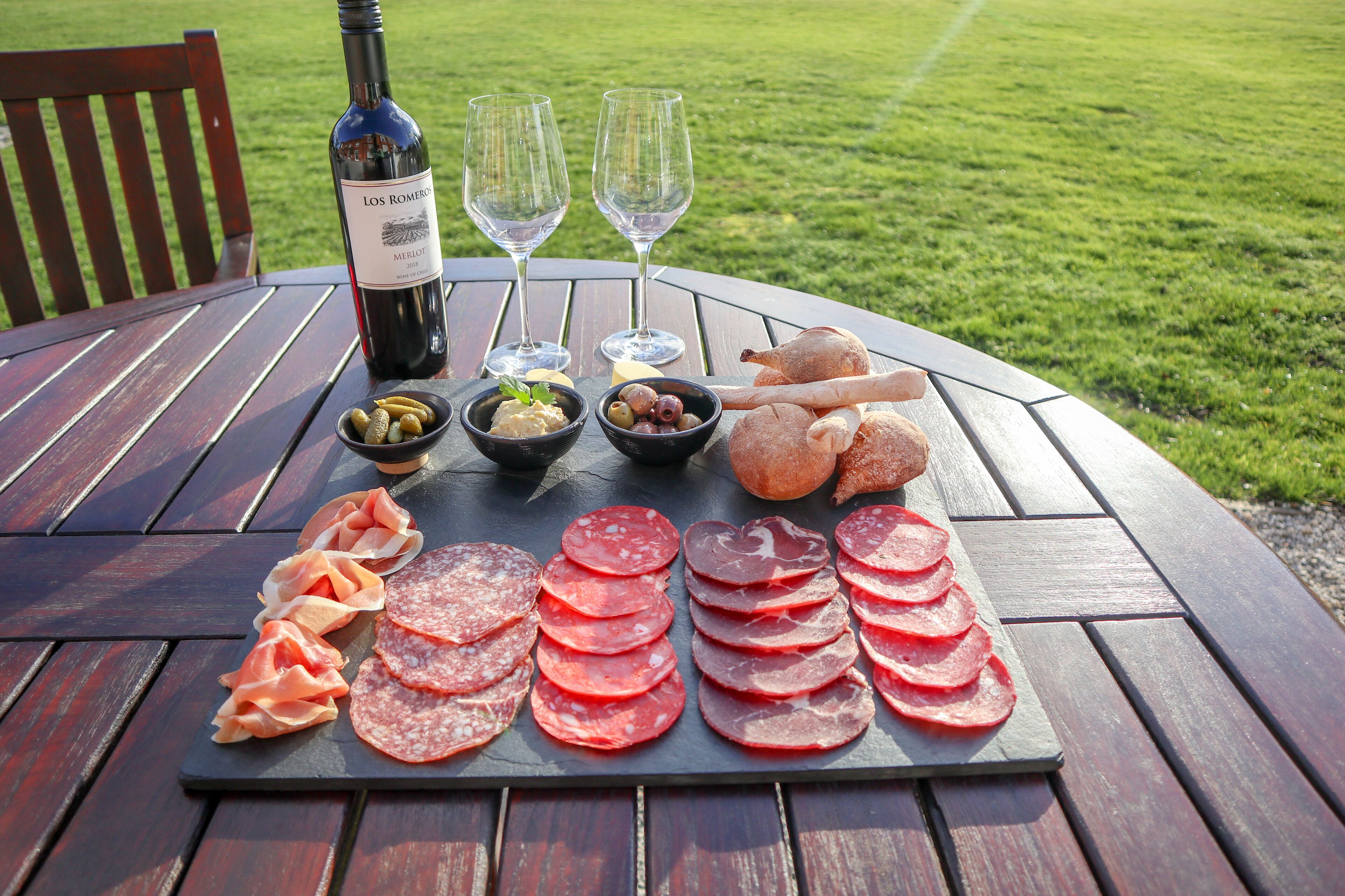 Une table avec une bouteille de vin, deux verres et un assortiment de nourriture comprenant du salami, des olives et du pain.