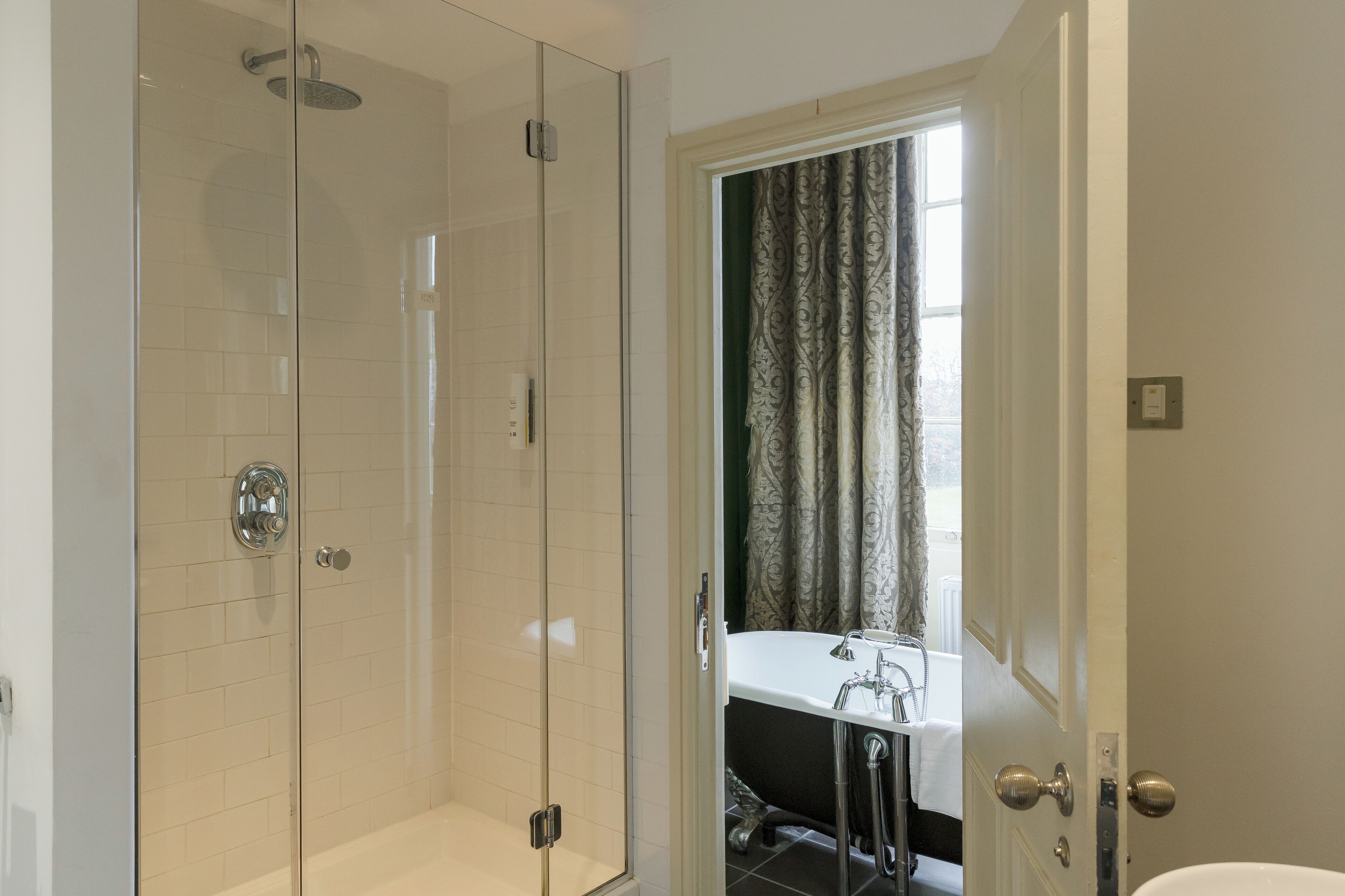 A modern bathroom with a glass-enclosed shower, a white bathtub, and a window with curtains.