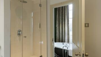A modern bathroom with a glass-enclosed shower, a white bathtub, and a window with curtains.