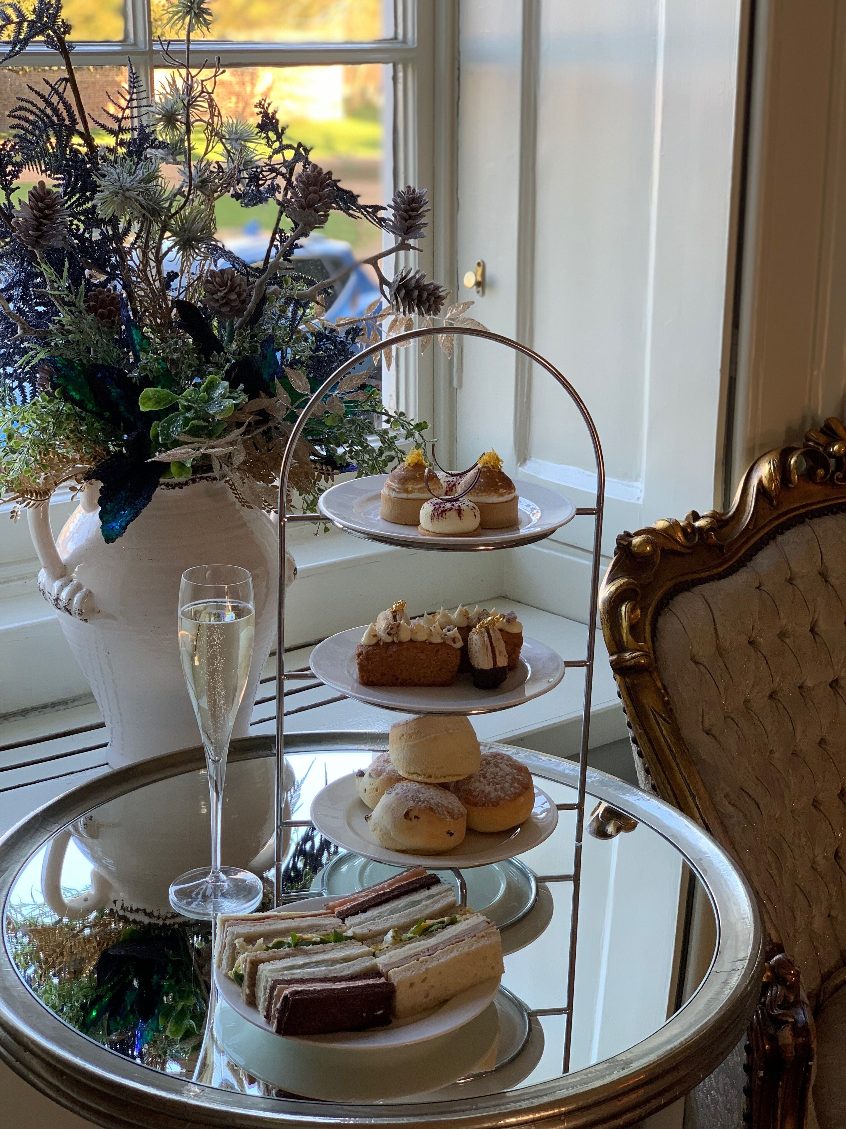 Un elegante soporte escalonado con pasteles y sándwiches, una copa de champán y una planta decorativa al fondo.