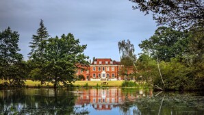 A large brick building with a symmetrical facade, a central entrance, and a reflection in the water.