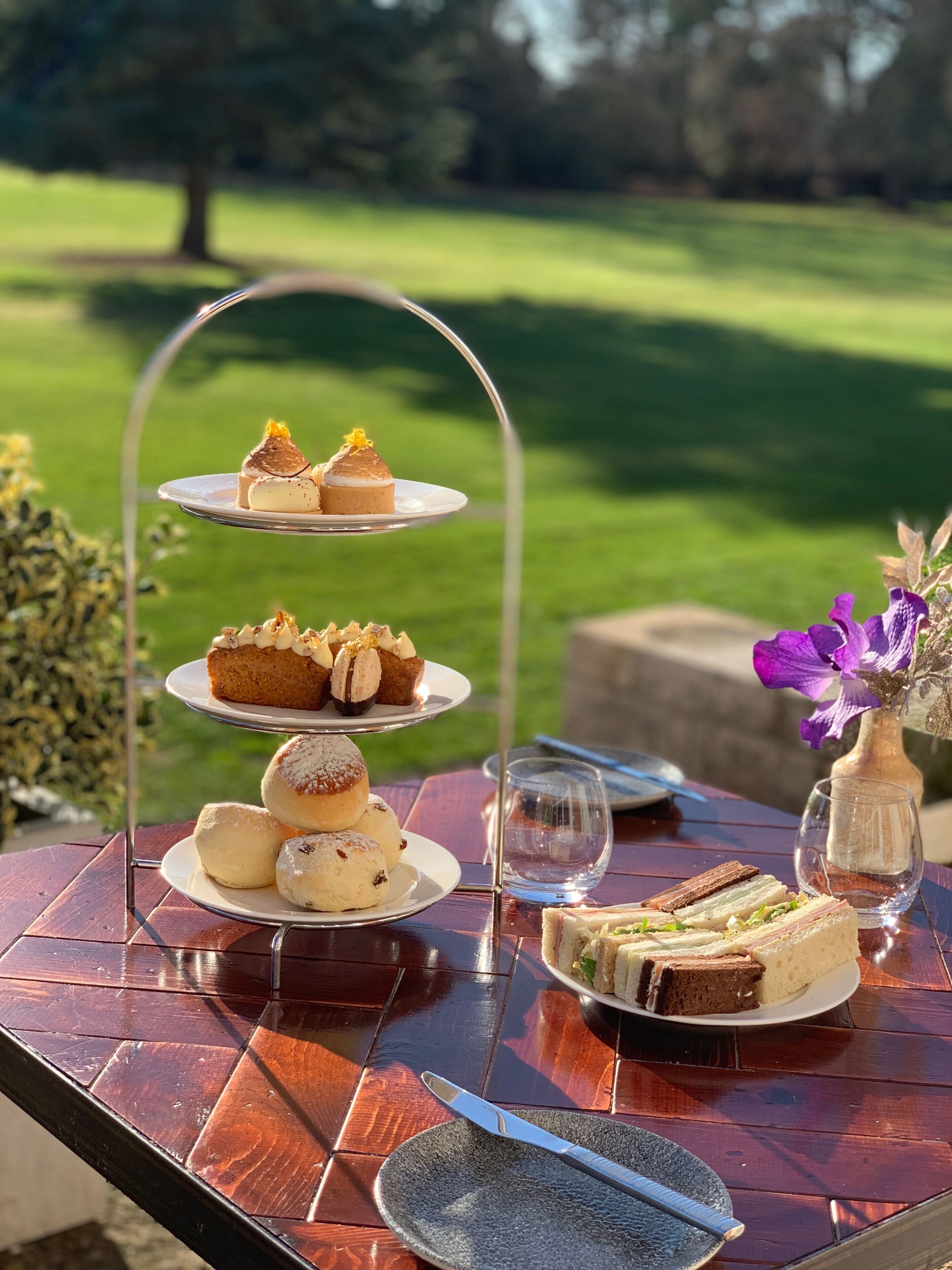 A tiered dessert stand with pastries and sandwiches on a wooden table outdoors.