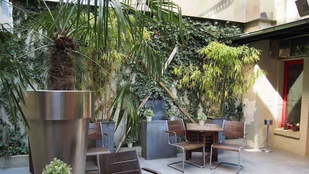 Un patio al aire libre con mesas y sillas de madera, rodeado de plantas en macetas y una jardinera de metal.