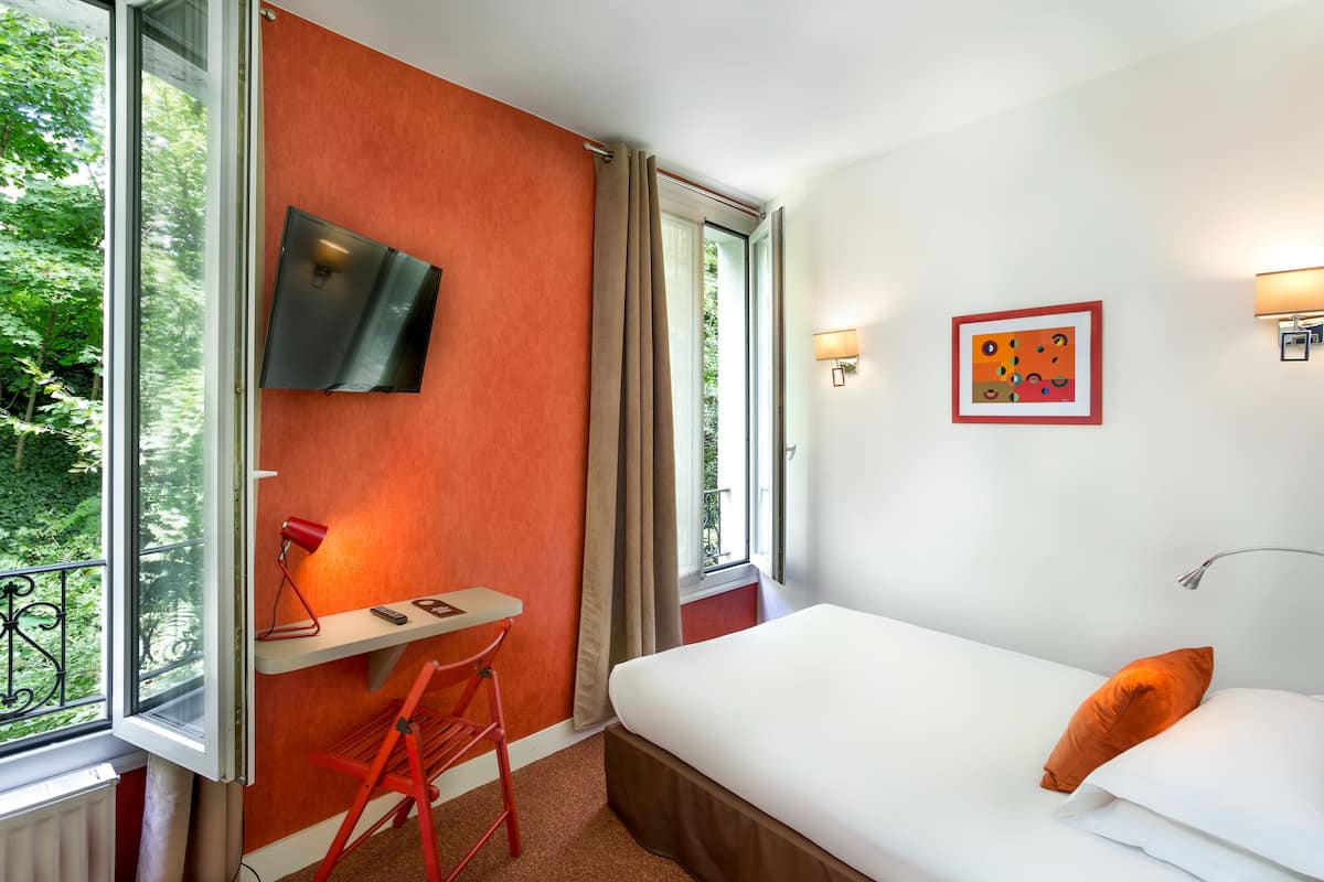 A hotel room with a bed, a desk, a TV, and a window with a view of greenery.