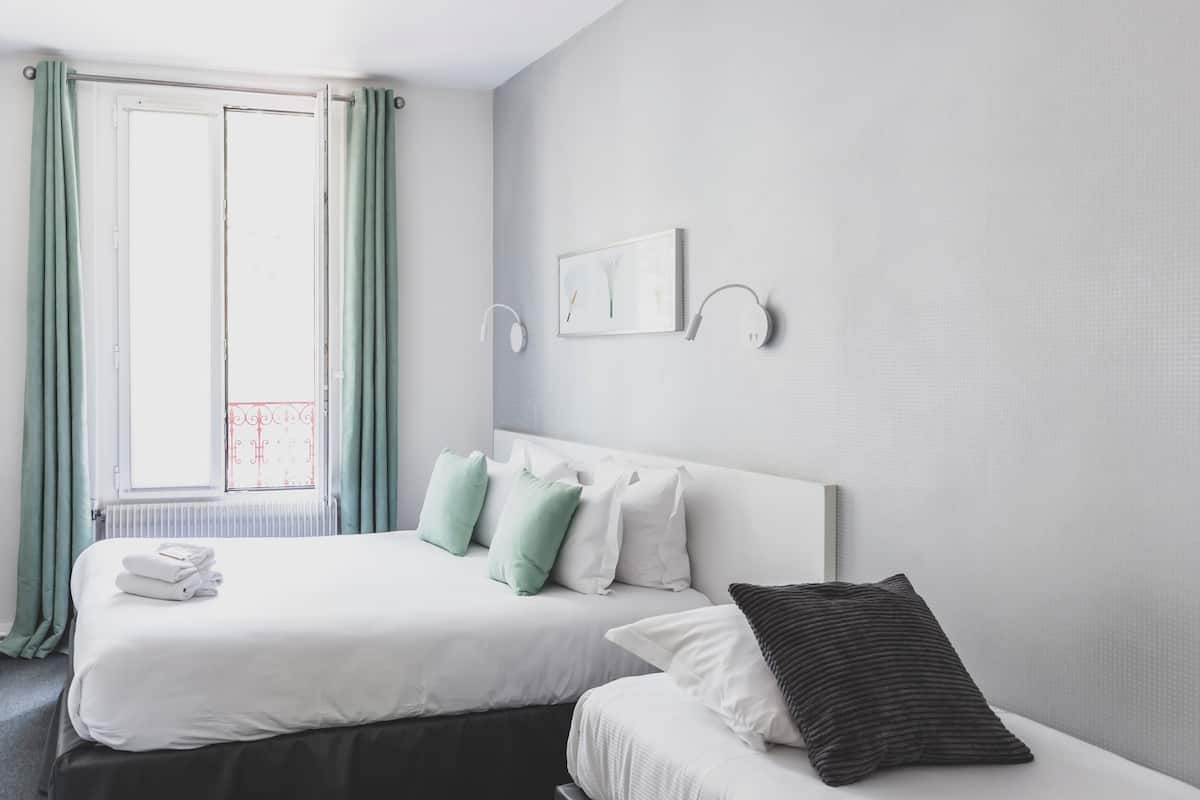 A hotel room with two beds, a window with curtains, a radiator, and a wall-mounted light fixture.