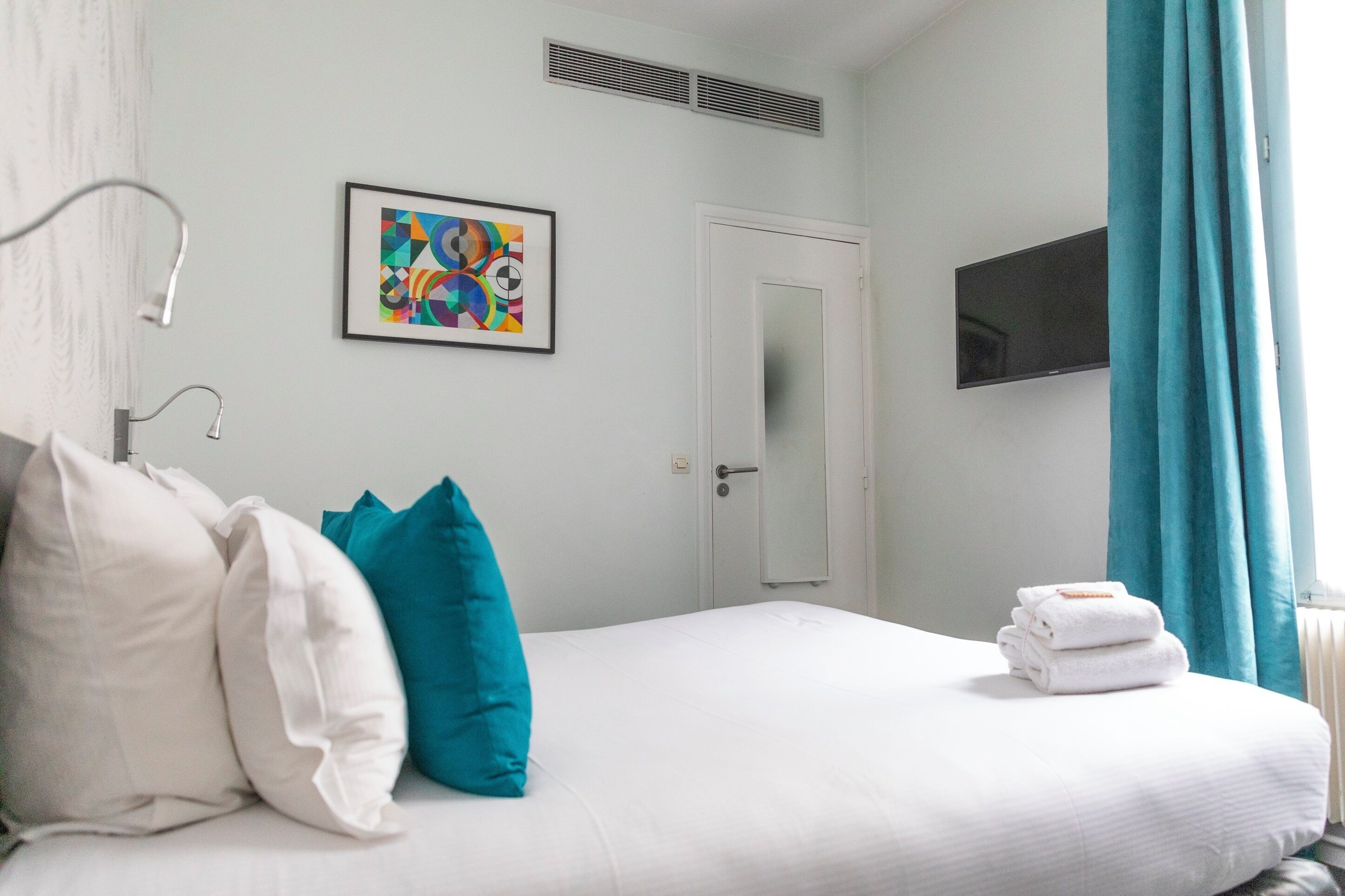 Une chambre d’hôtel avec un lit, des oreillers, un tableau abstrait encadré, une télévision et une fenêtre avec des rideaux.