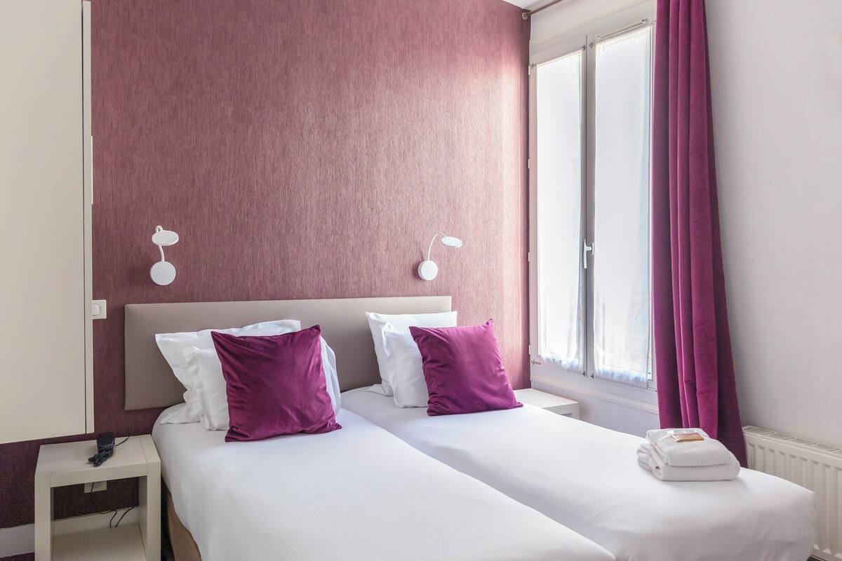 A double bed with white and purple pillows, a white bedside table, and a window with purple curtains.