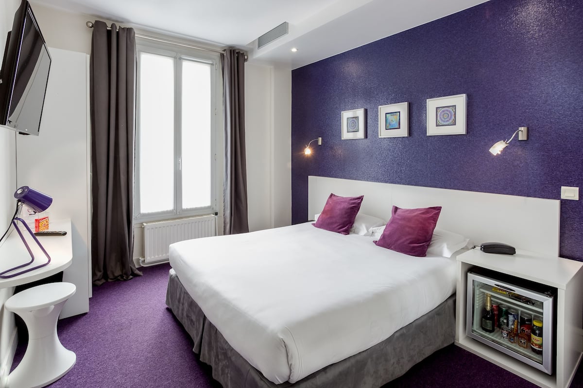 A hotel room with a bed, a desk, a chair, a TV, a window with curtains, and wall-mounted lights.