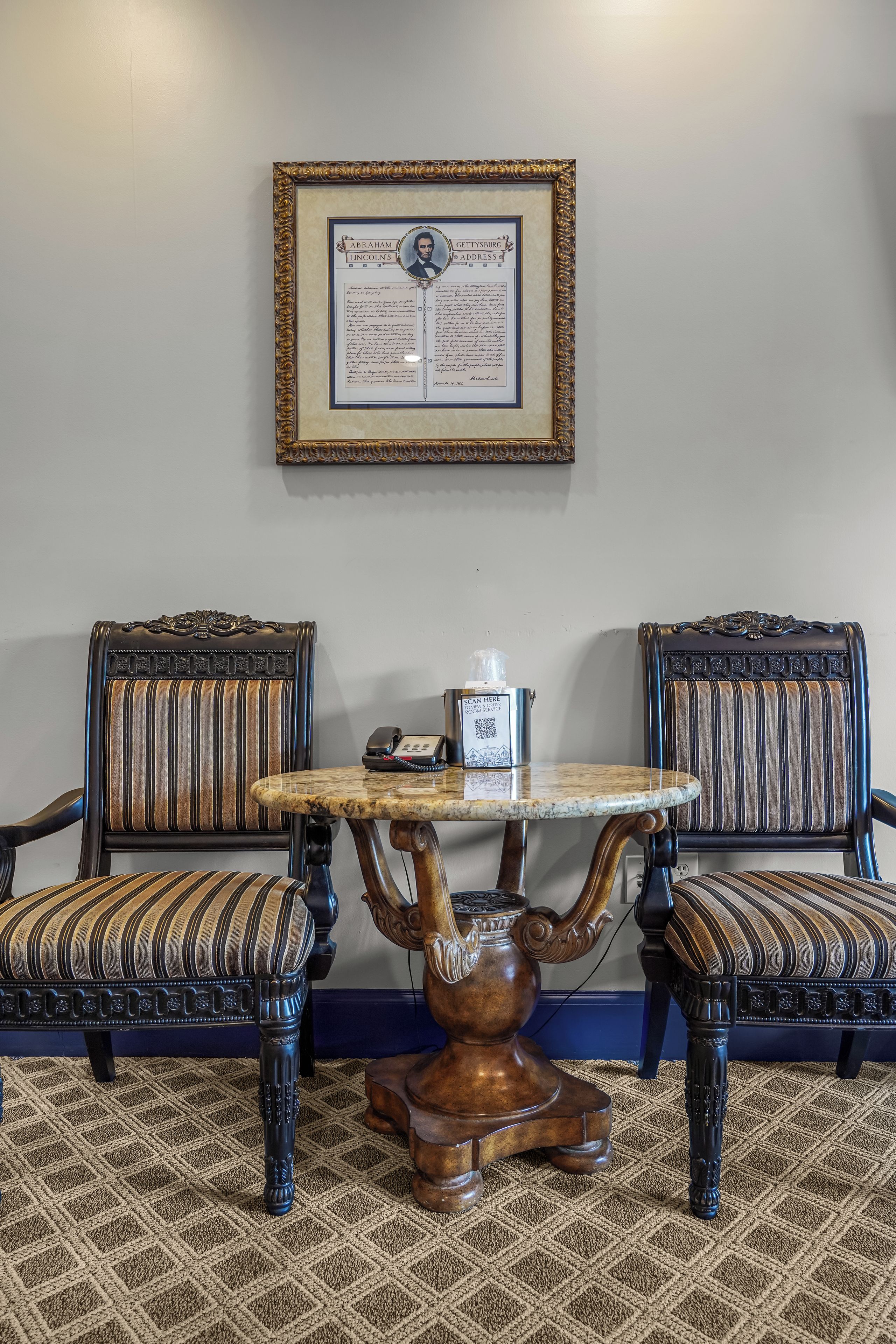 Een kamer met twee gestoffeerde fauteuils, een klein rond tafeltje met een telefoon, en een ingelijst historisch document aan de muur.
