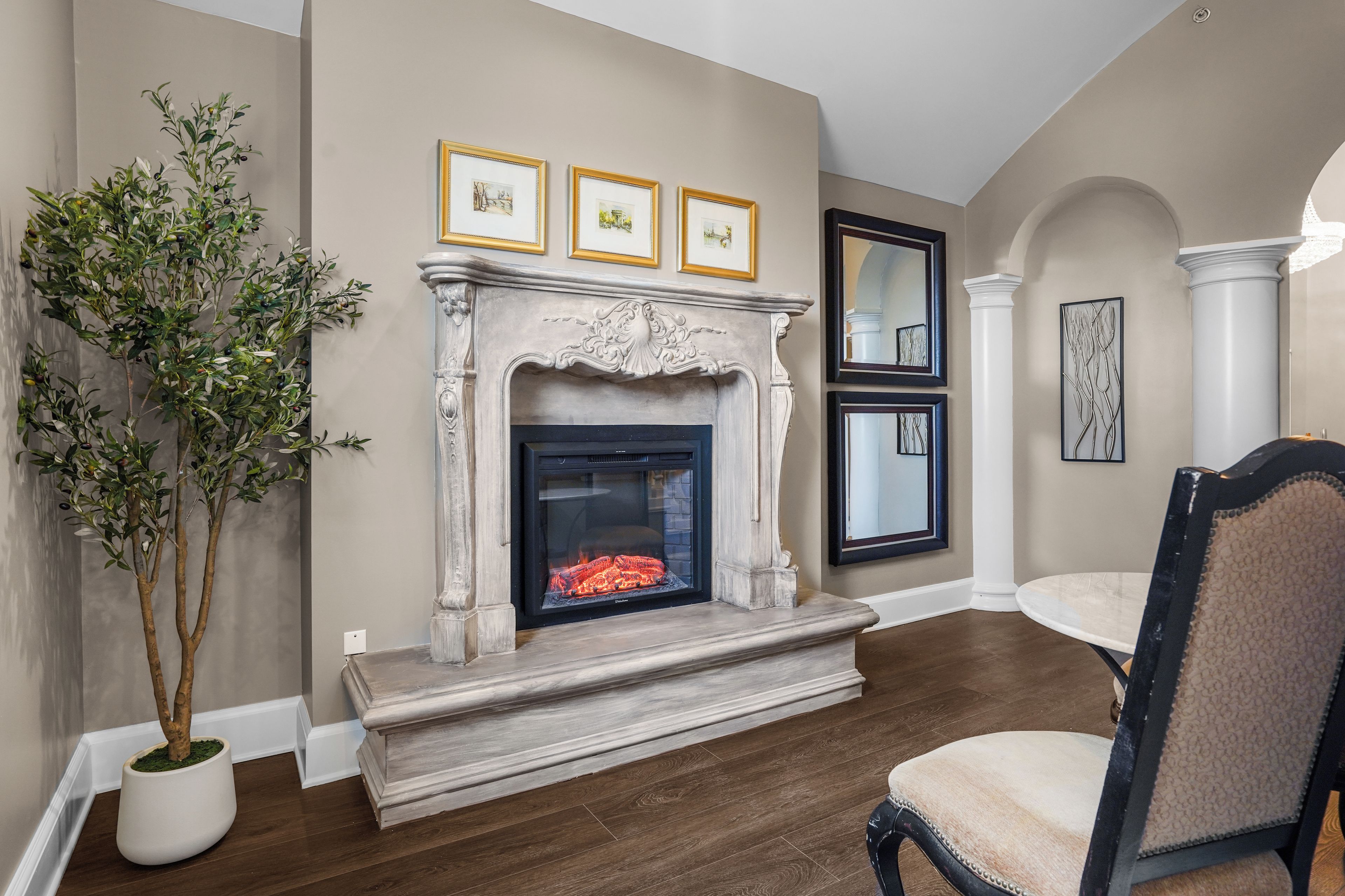 A room with a fireplace, framed pictures on the wall, a potted plant, and a dining area with a chair.