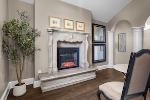 A room with a fireplace, framed pictures on the wall, a potted plant, and a dining area with a chair. - Chateau Avalon Hotel & Spa (Kansas City)