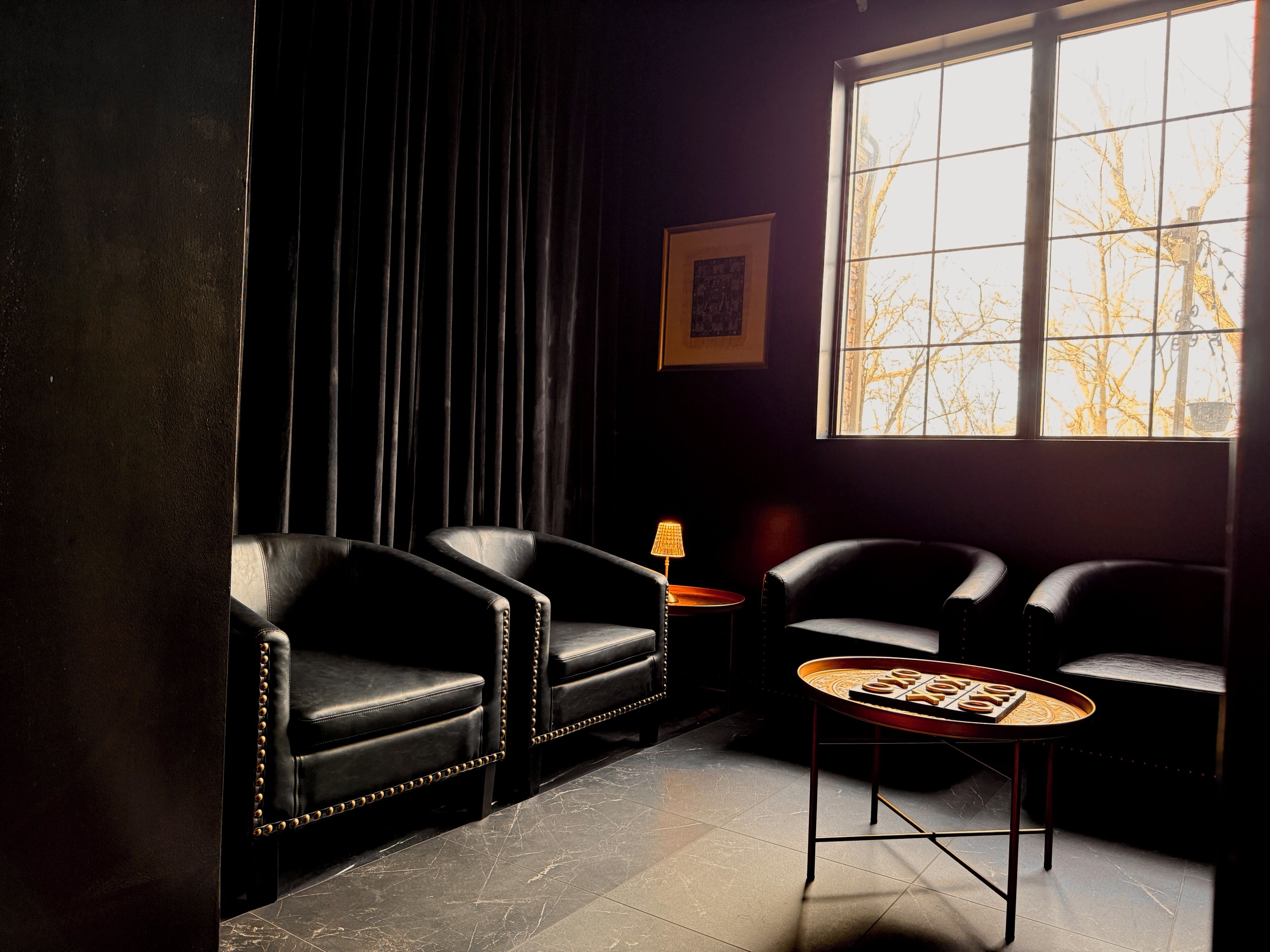 Una habitación con dos sillones de cuero, una mesita con una lámpara y un ventanal con vistas a árboles desnudos.