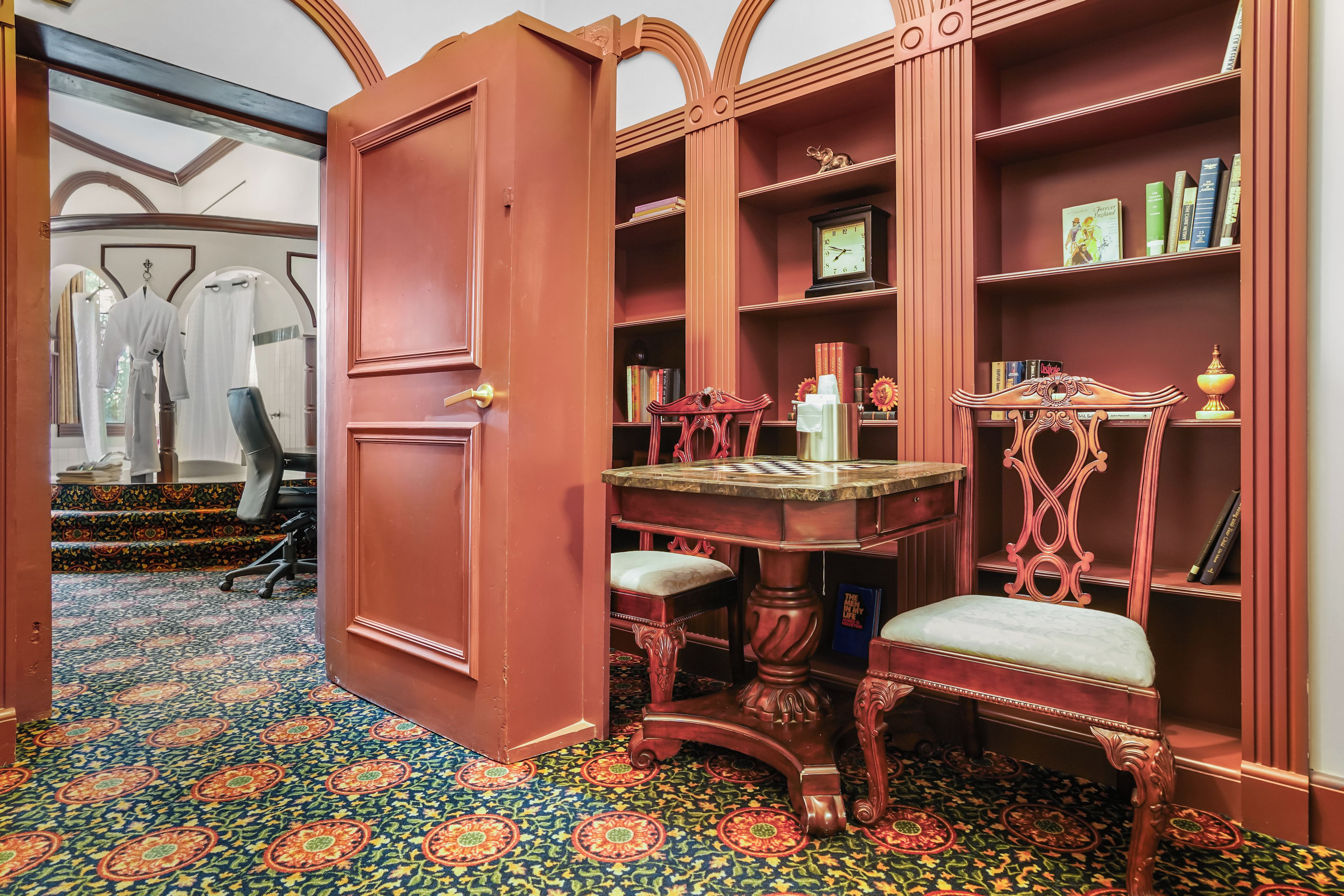 Una habitación con una gran puerta de madera, una mesa con cubierta de mármol, una silla tapizada en rojo y una estantería con libros y objetos decorativos.