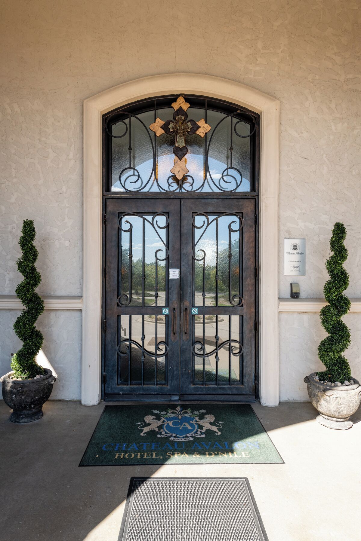 A double door with a decorative crest, a doormat with hotel name, and two potted plants on either side.