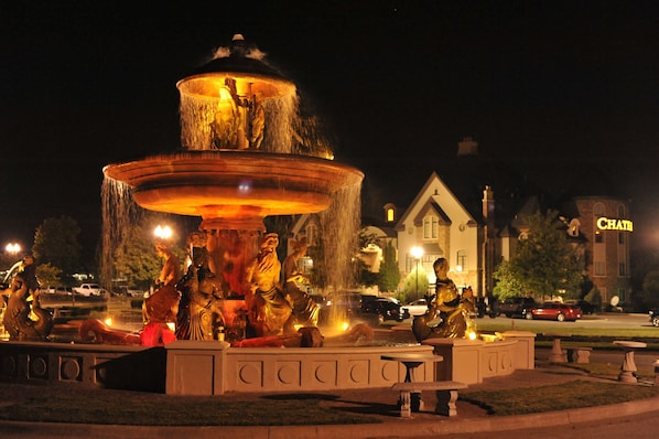 A multi-tiered fountain with statues, surrounded by a paved area with benches, set against a night-time urban backdrop. - Chateau Avalon Hotel & Spa (Kansas City)
