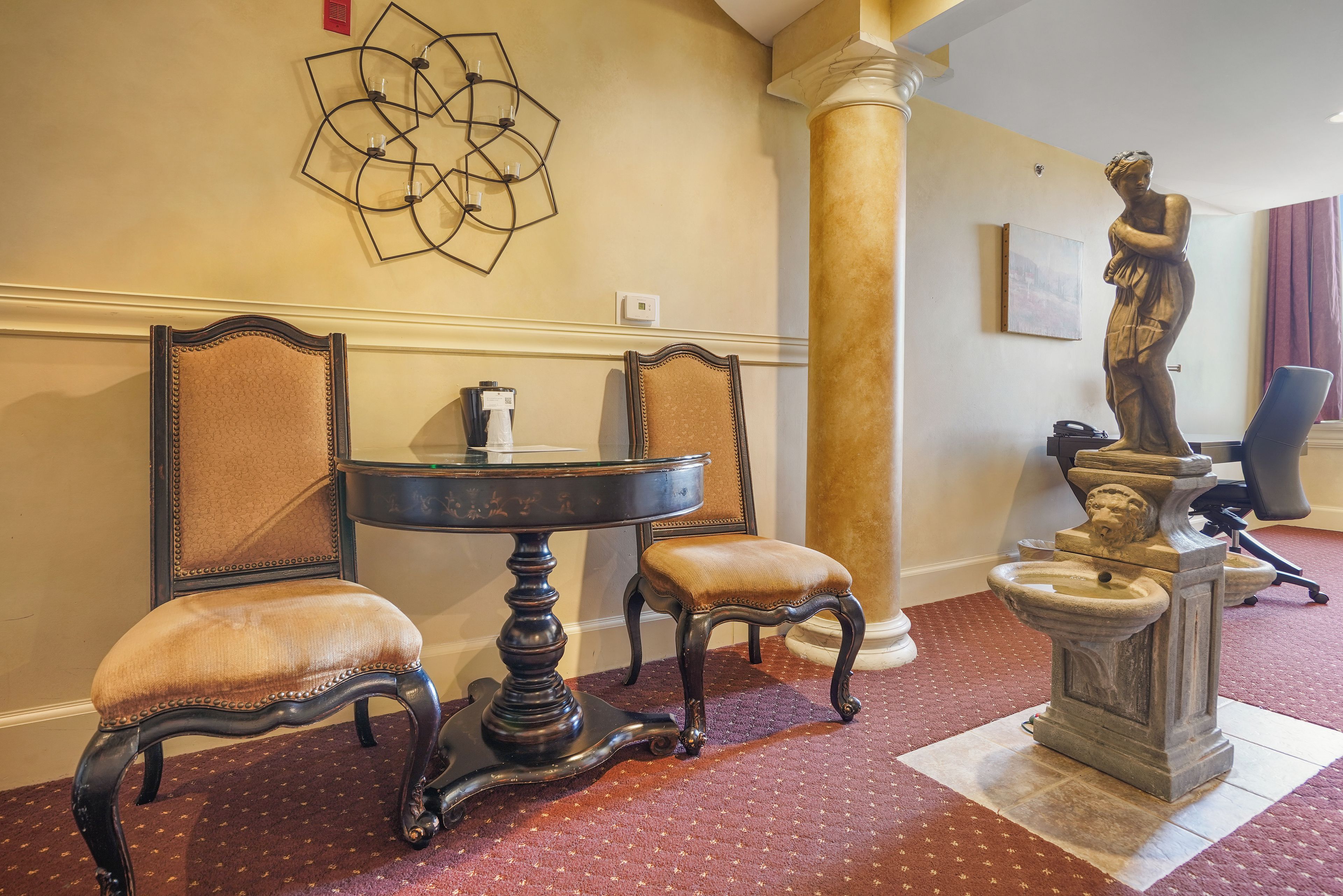 A room with a round table, two chairs, a statue, and a wall-mounted abstract art piece.