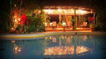 A pool area with a covered seating area, surrounded by potted plants and trees.