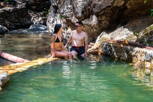 Una pareja sentada junto a una piscina cristalina, rocosa, con un muro de piedra y una tubería.